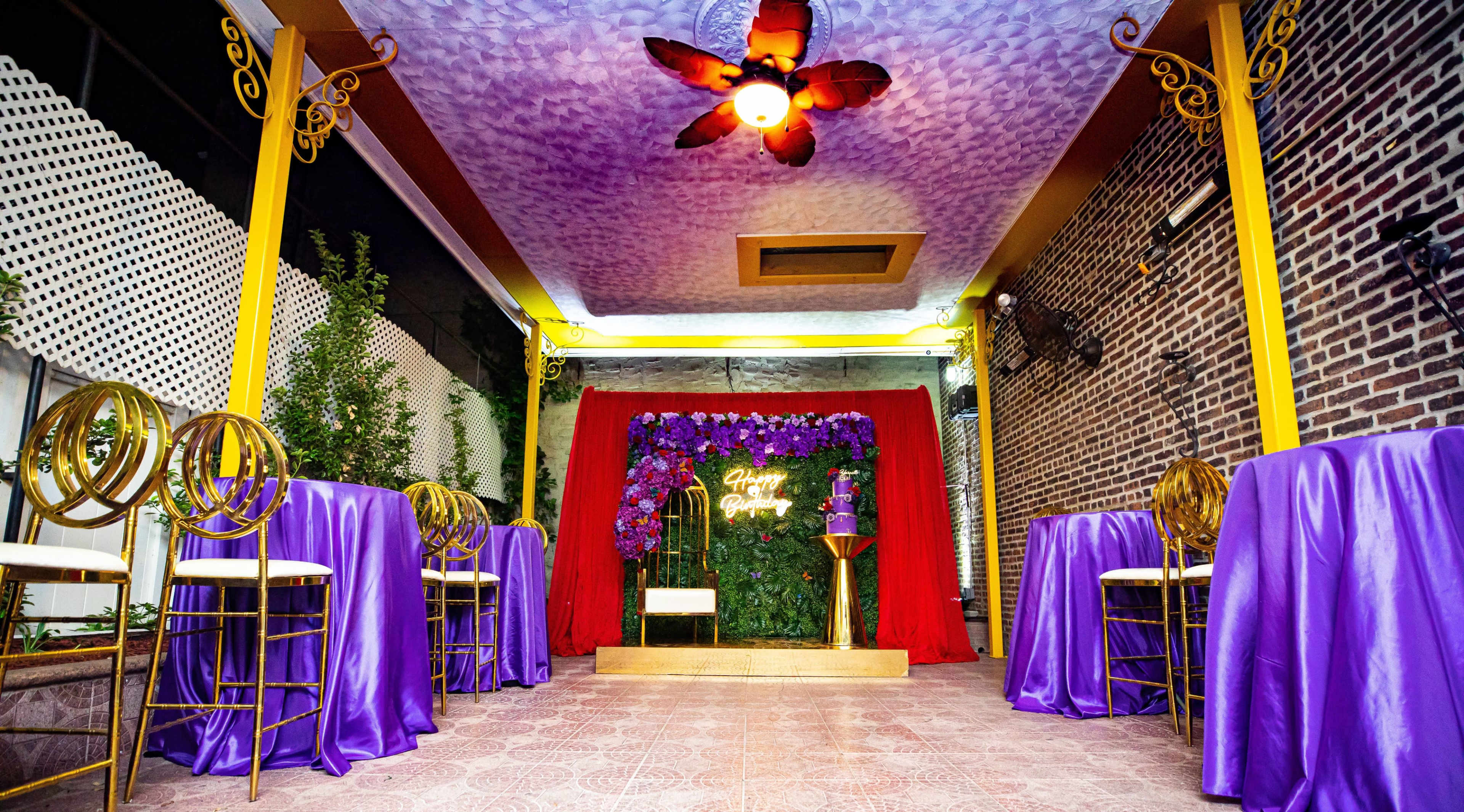 The image shows an event space decorated with purple tablecloths, gold accents, and a floral backdrop for a celebration.