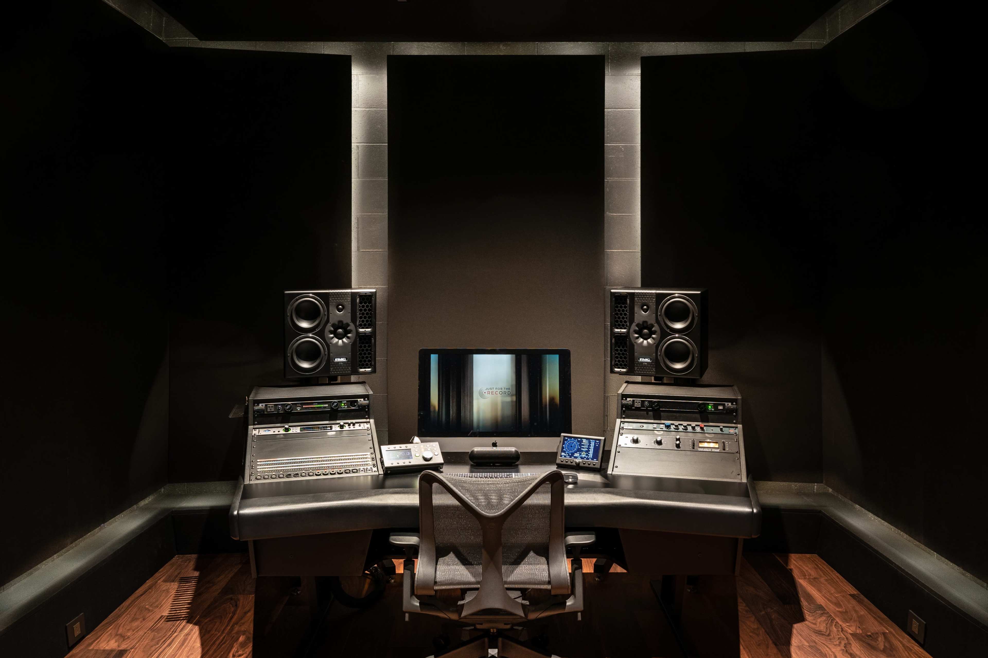 The image shows a music production studio with a central workstation, two large speakers, and a computer monitor.