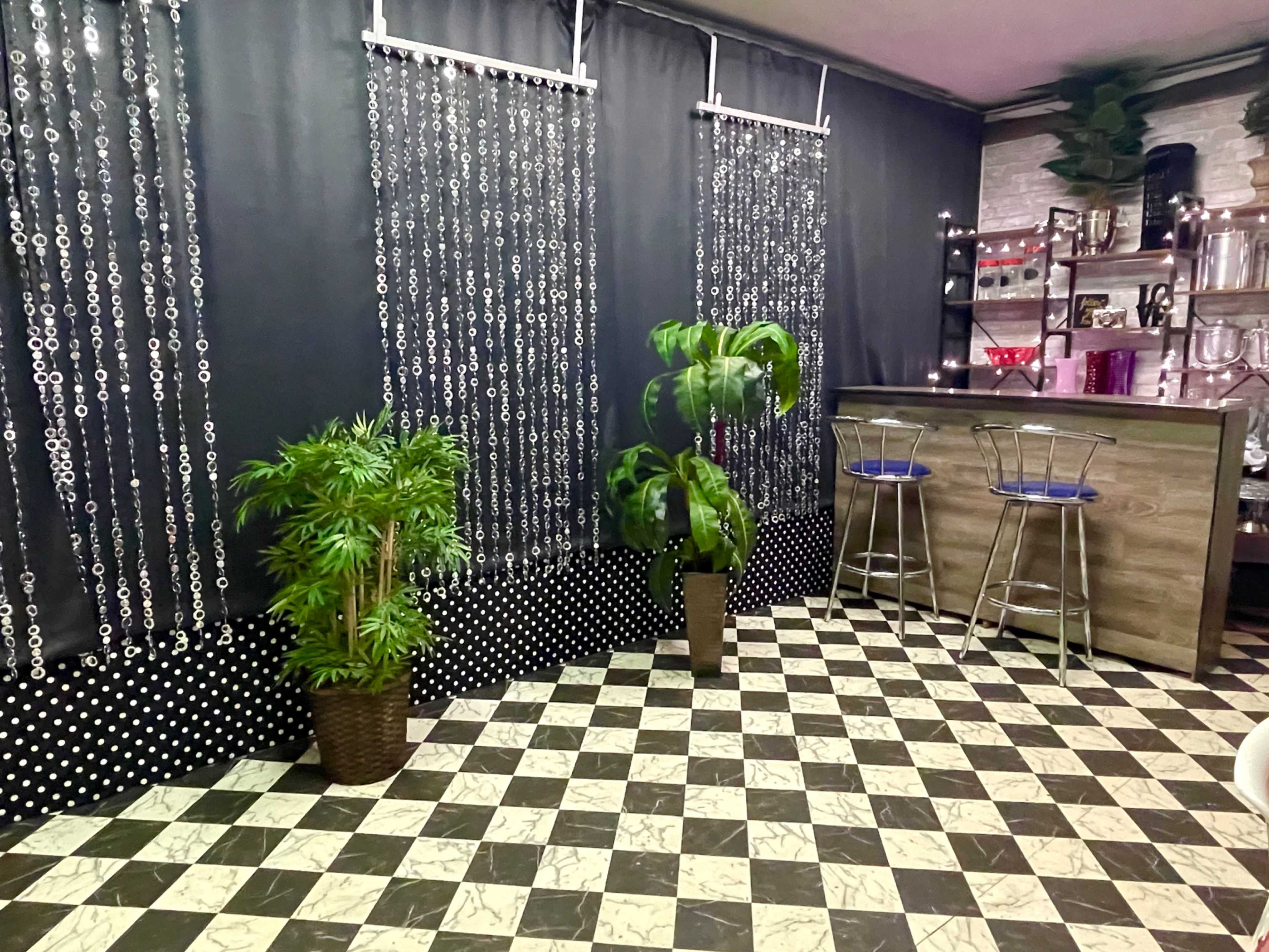 A room with a checkered floor, a bar area, and two potted plants beside a black curtain adorned with hanging beads.
