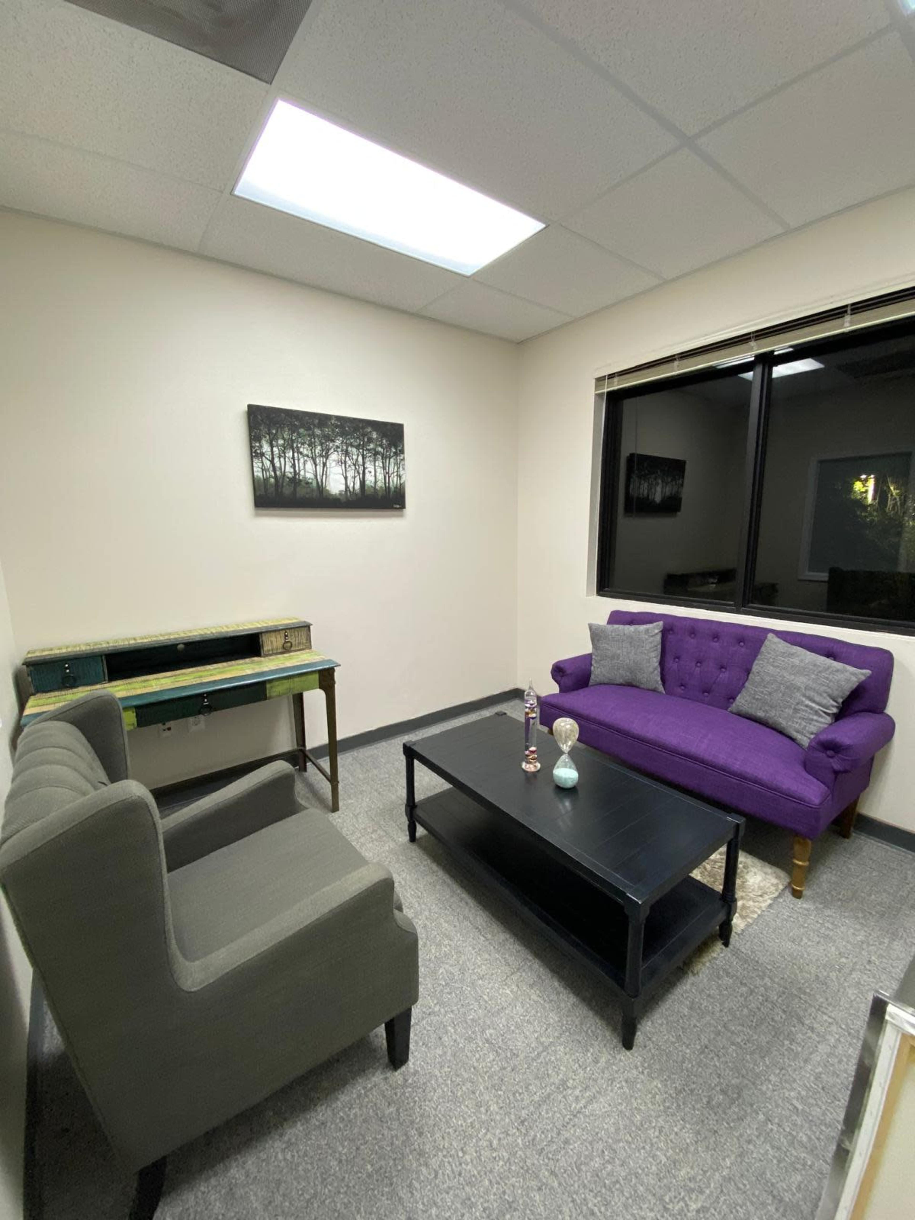 The image shows a small, well-lit office lounge area with a purple sofa, two gray armchairs, a black coffee table, and a large window.