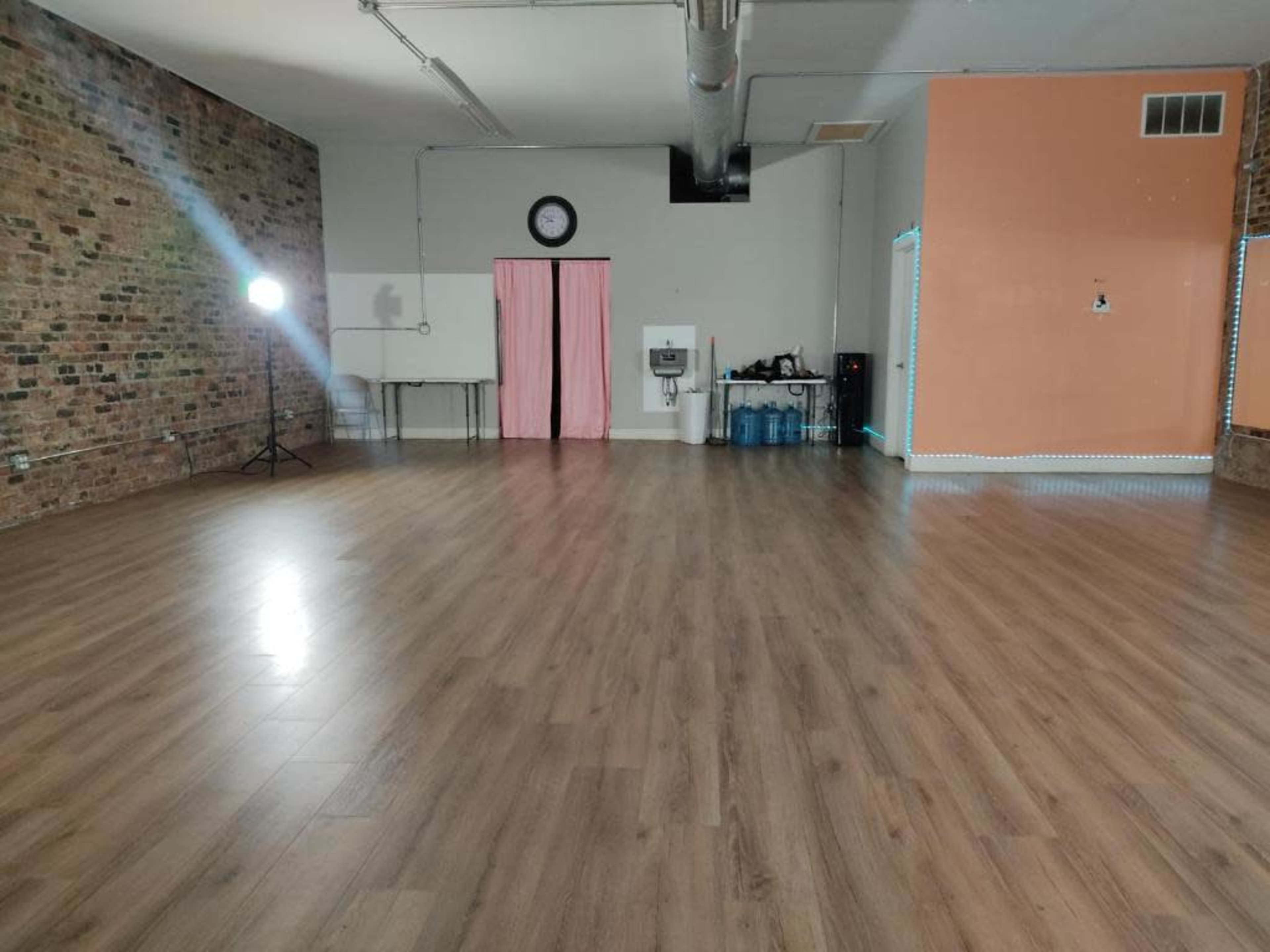 A spacious room with wooden floors, exposed brick walls, and minimal furniture, featuring a clock on the wall and a pink curtain.