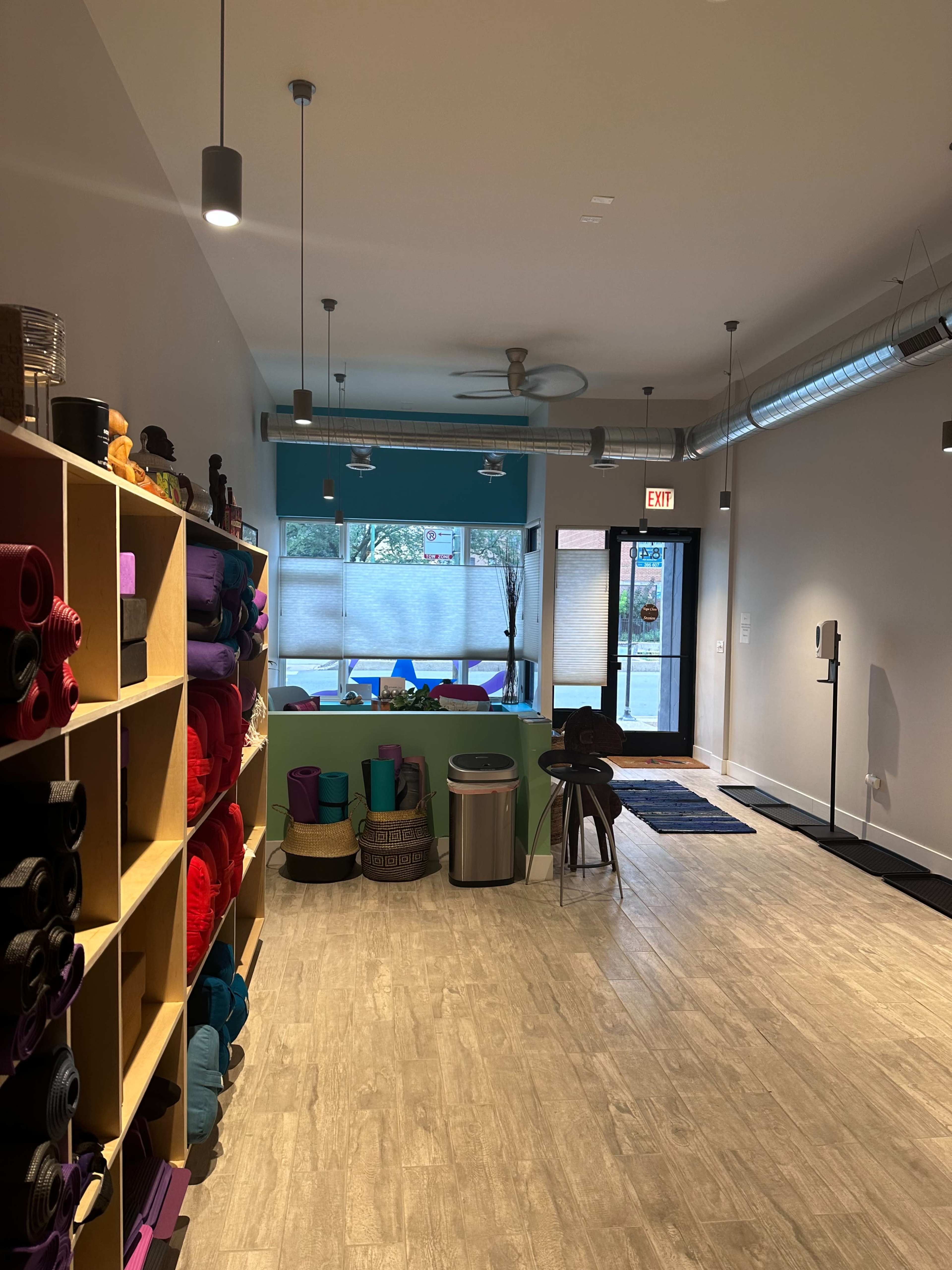 The image shows a yoga studio interior with neatly arranged shelves of colorful yoga mats on the left and a view of large windows with partially drawn shades on the right.