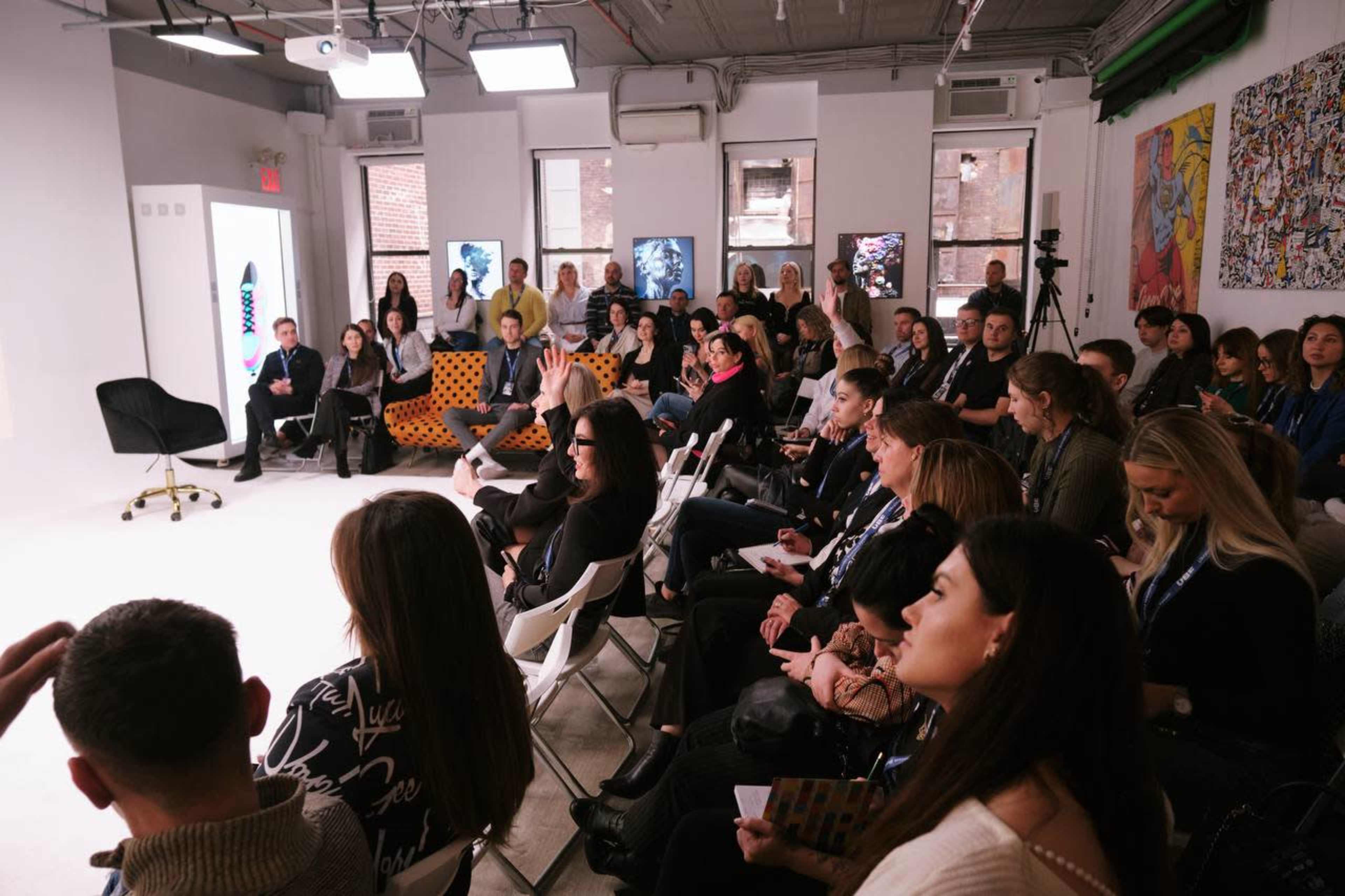 A group of people sits in a well-lit room, attentively facing a speaker at the front with art on the walls and chairs arranged in rows.