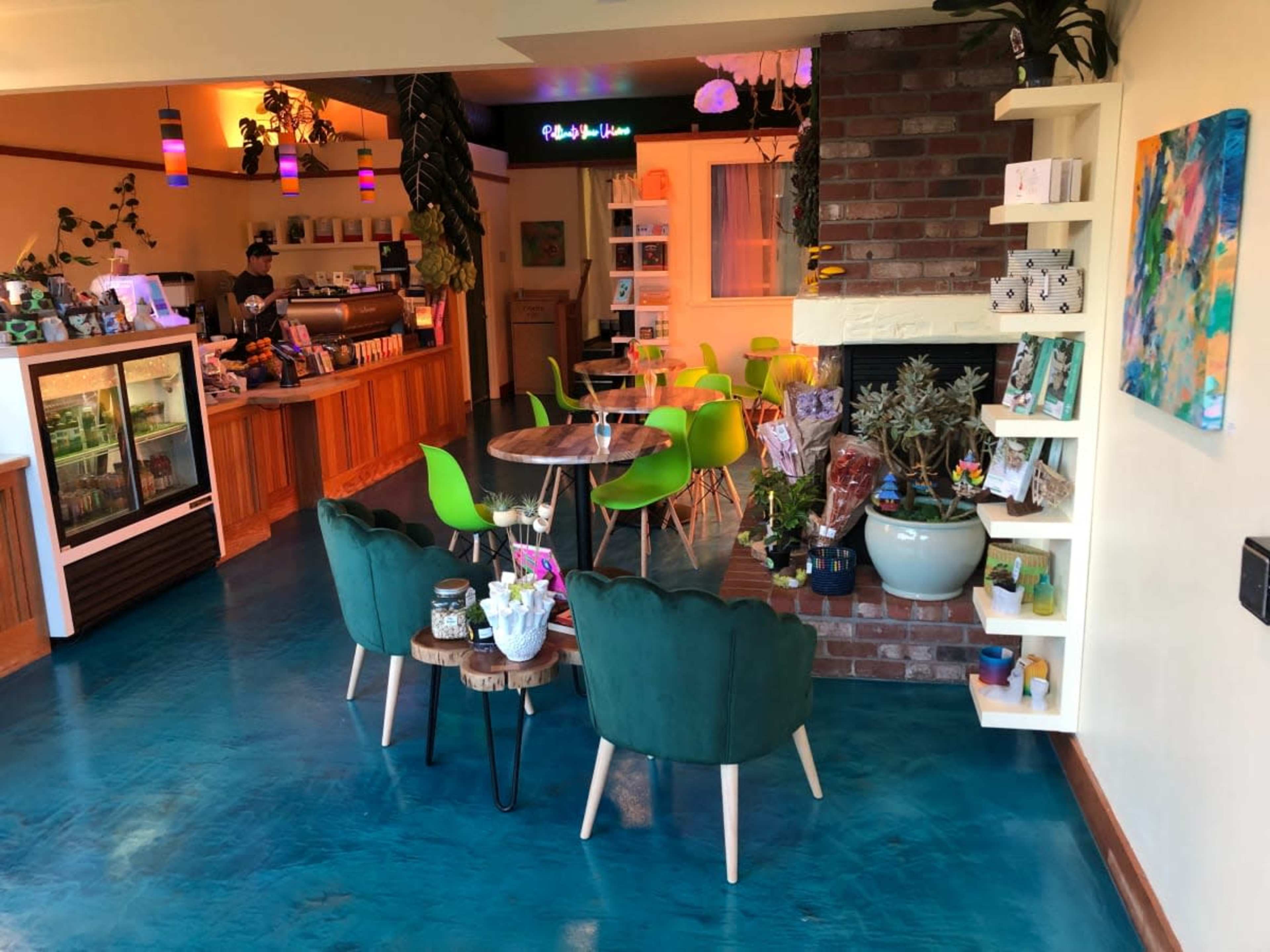 The image shows a modern cafe interior with green chairs, wooden tables, and a decorative wall featuring plants and artwork.