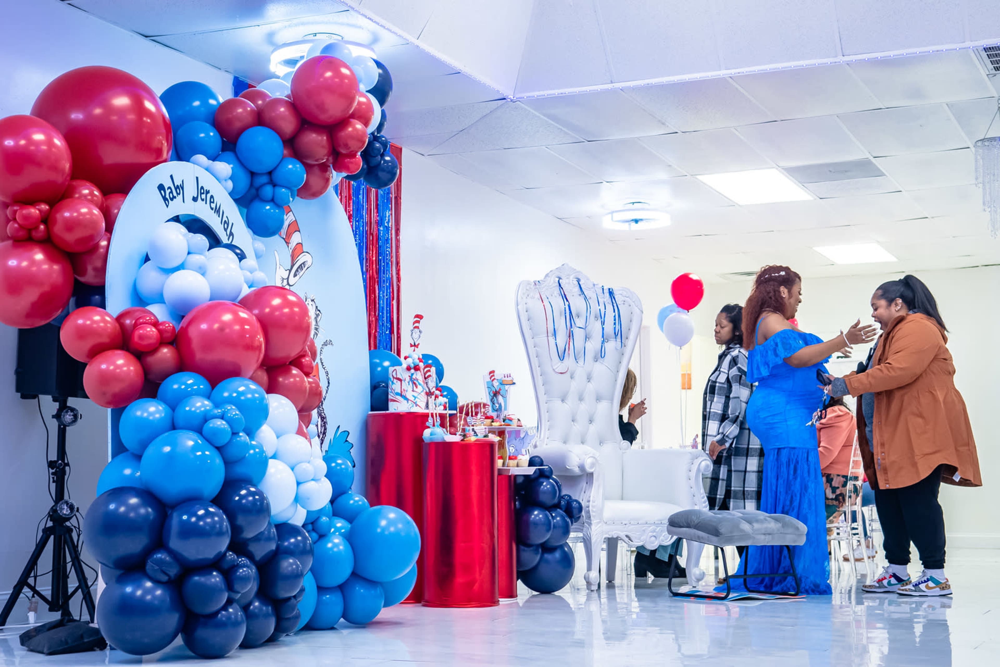A decorated event space features a colorful balloon arch, a cake table, and guests engaged in conversation.