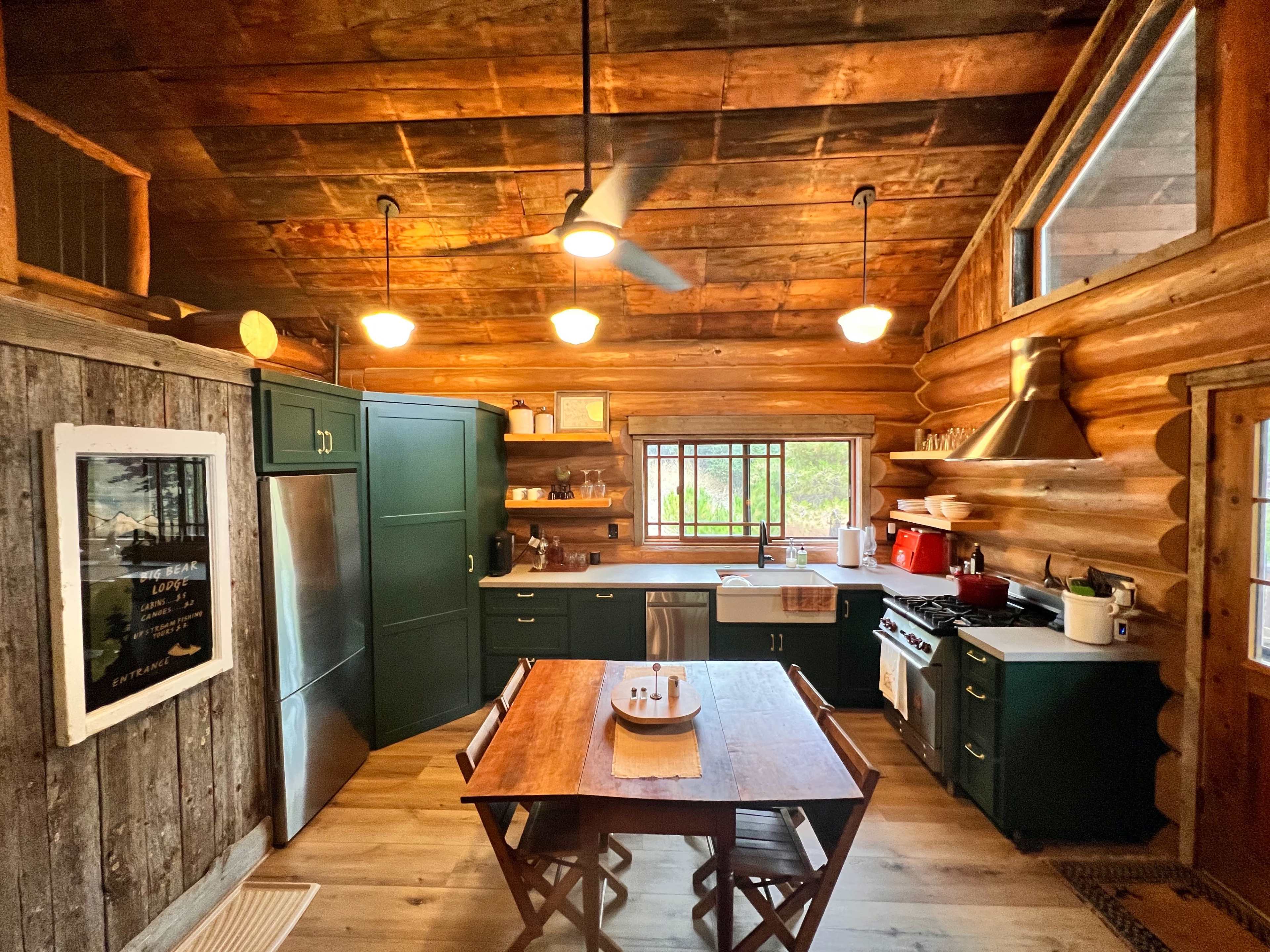 A cozy kitchen features a wooden log interior, green cabinetry, a dining table, and a large window providing natural light.