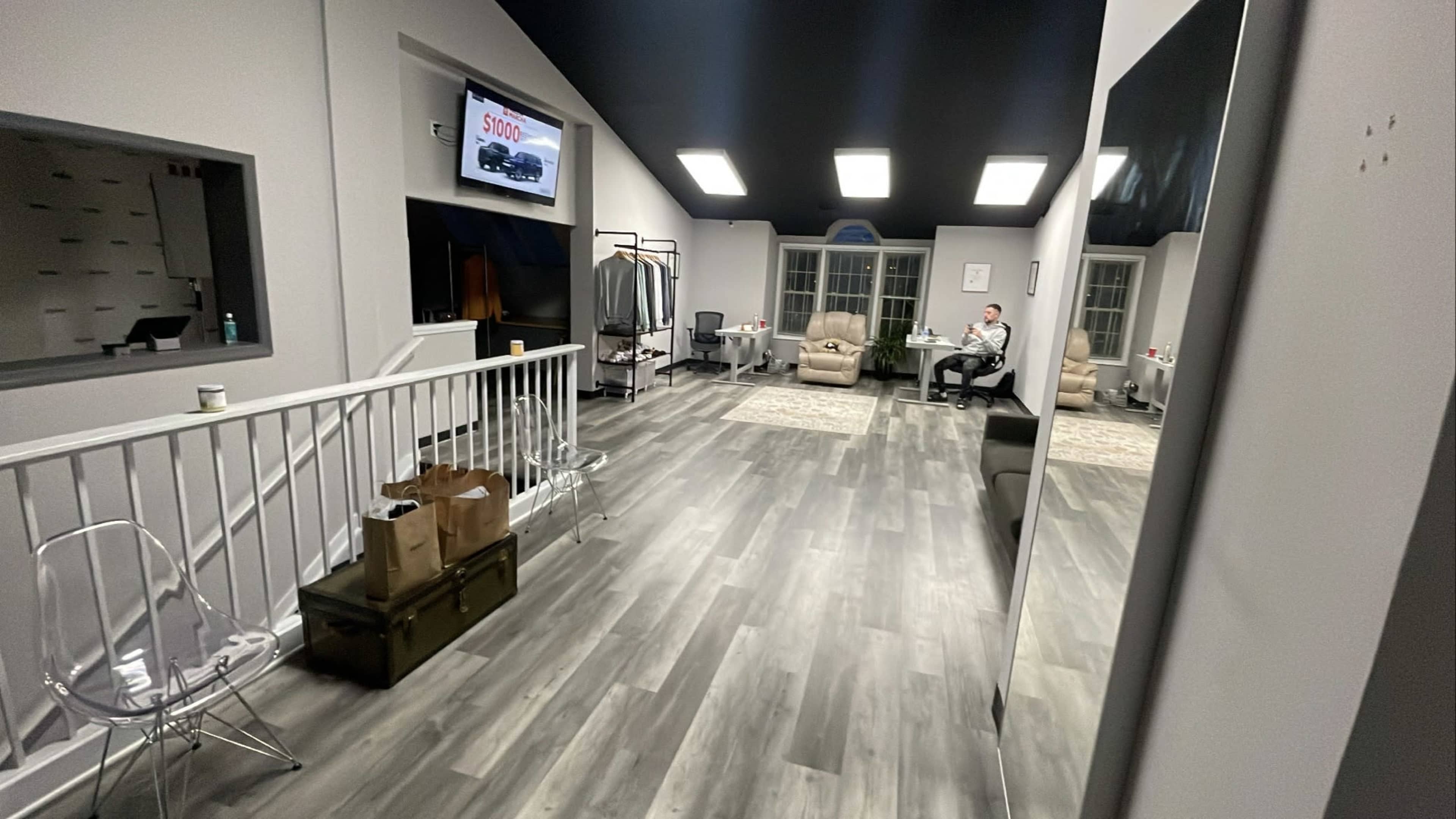 A spacious room with a modern design, featuring a seating area, a large mirror, and a television mounted on the wall, adjacent to a staircase and a window.