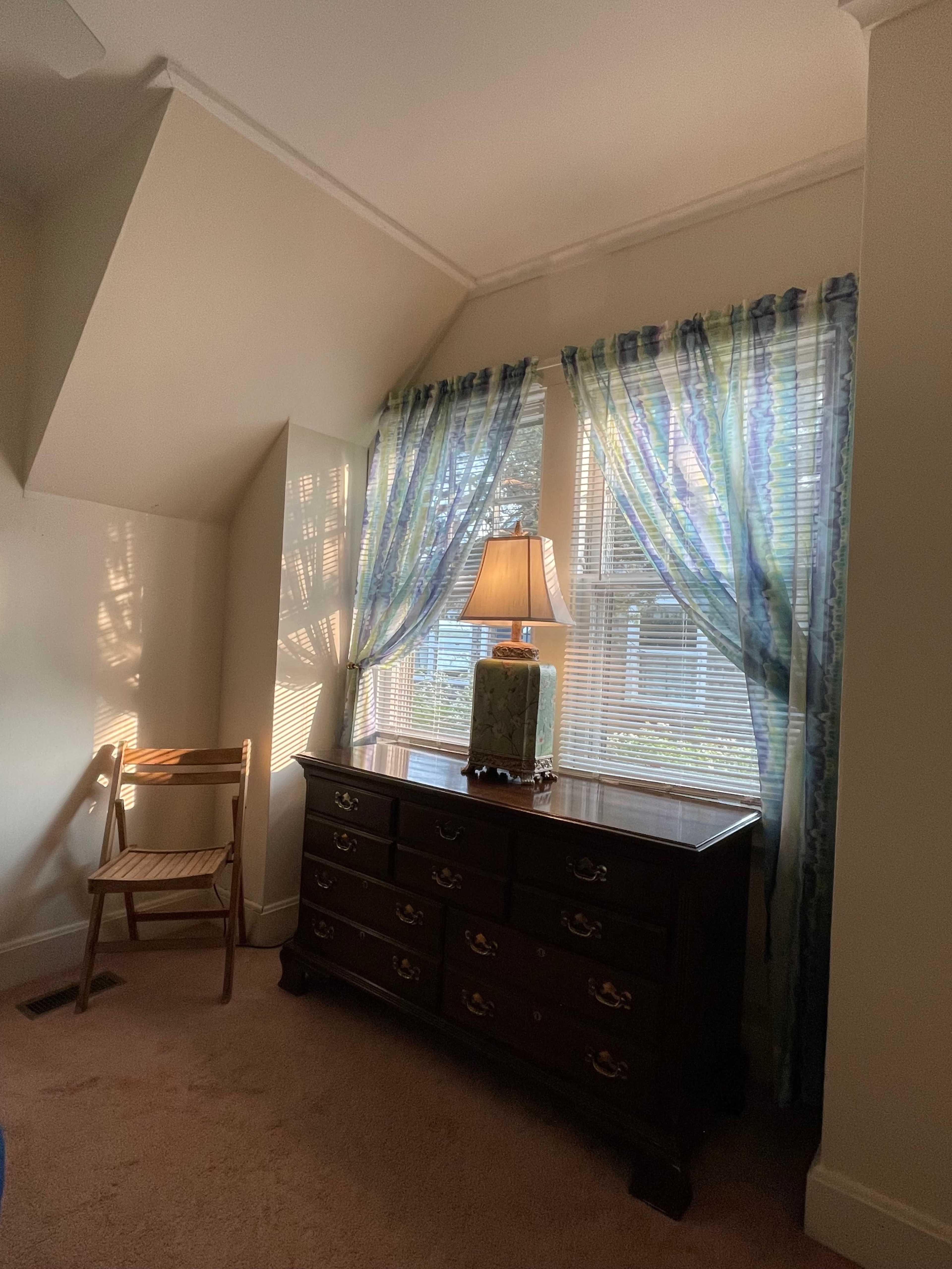 A wooden dresser with a lamp sits next to a chair in a well-lit corner of a room featuring light curtains over a window.