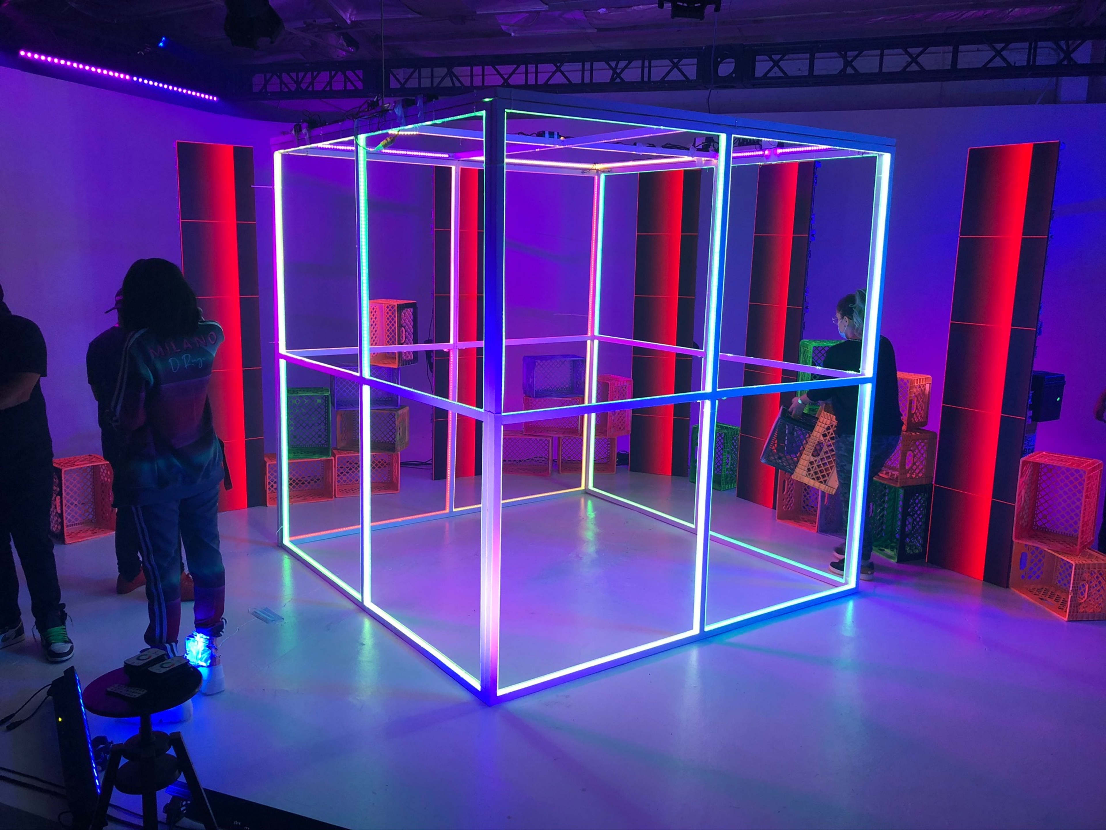 A brightly lit space features a geometric structure made of illuminated frames surrounded by colorful crates.