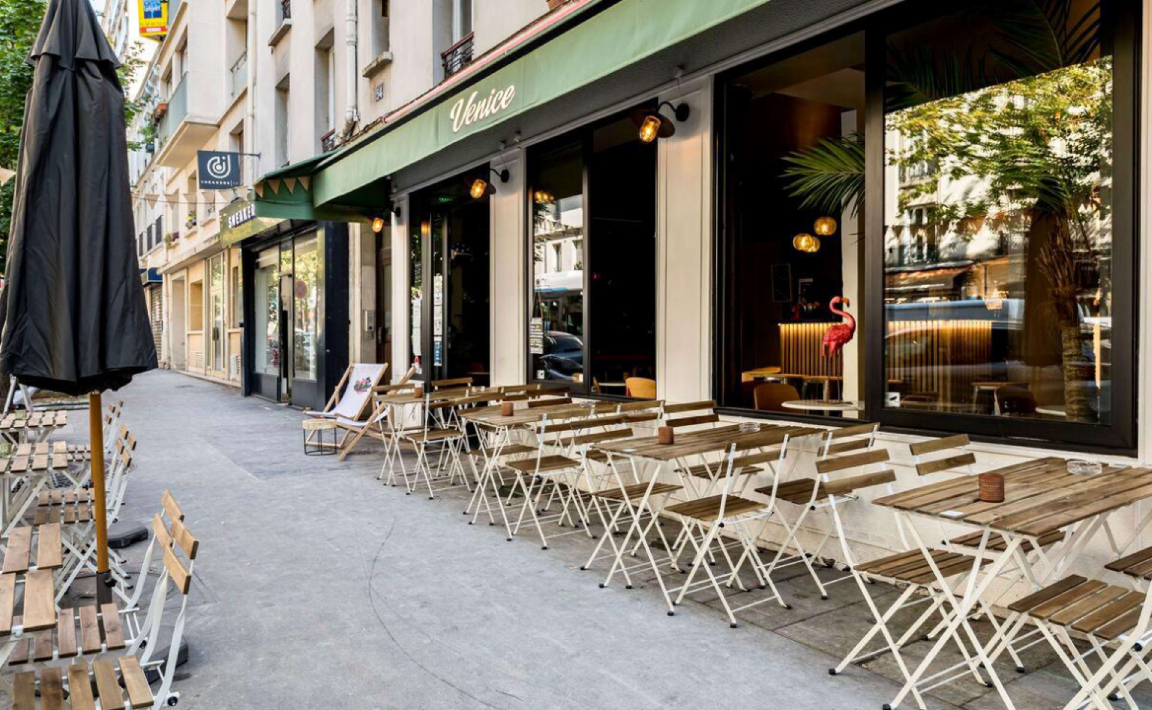 A restaurant patio with small wooden tables and chairs is situated on a sidewalk, next to large glass windows and a green awning.