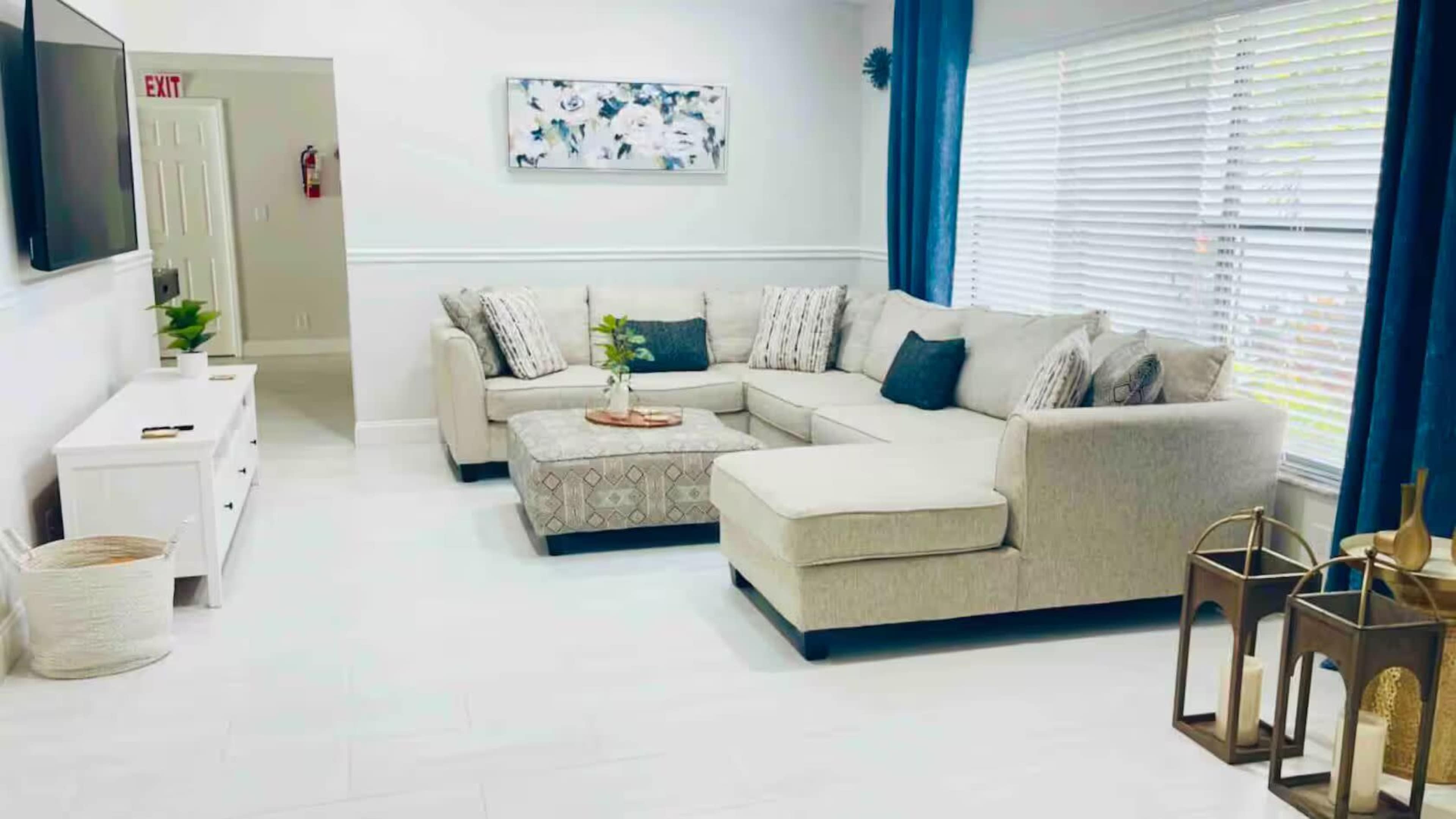 The image shows a neatly arranged living room with a light-colored sectional sofa, a coffee table, and decorative elements including a plant and lanterns.