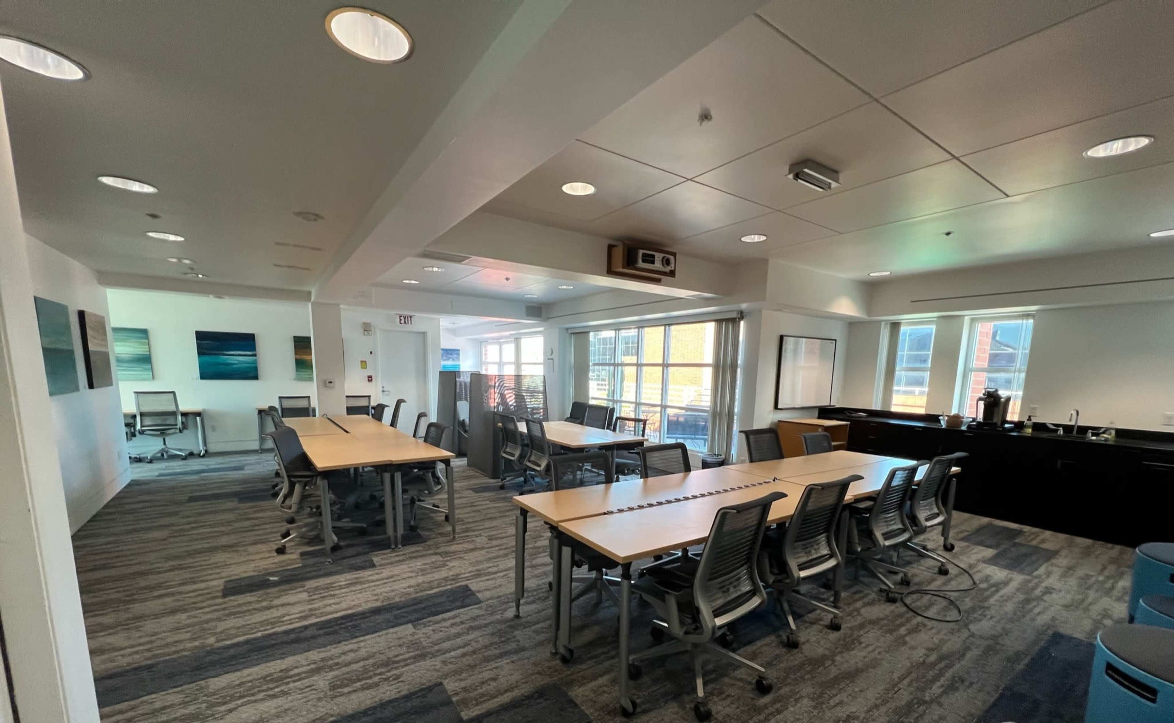 A modern conference room with multiple tables and chairs arranged around a central space, large windows, and a kitchenette area.