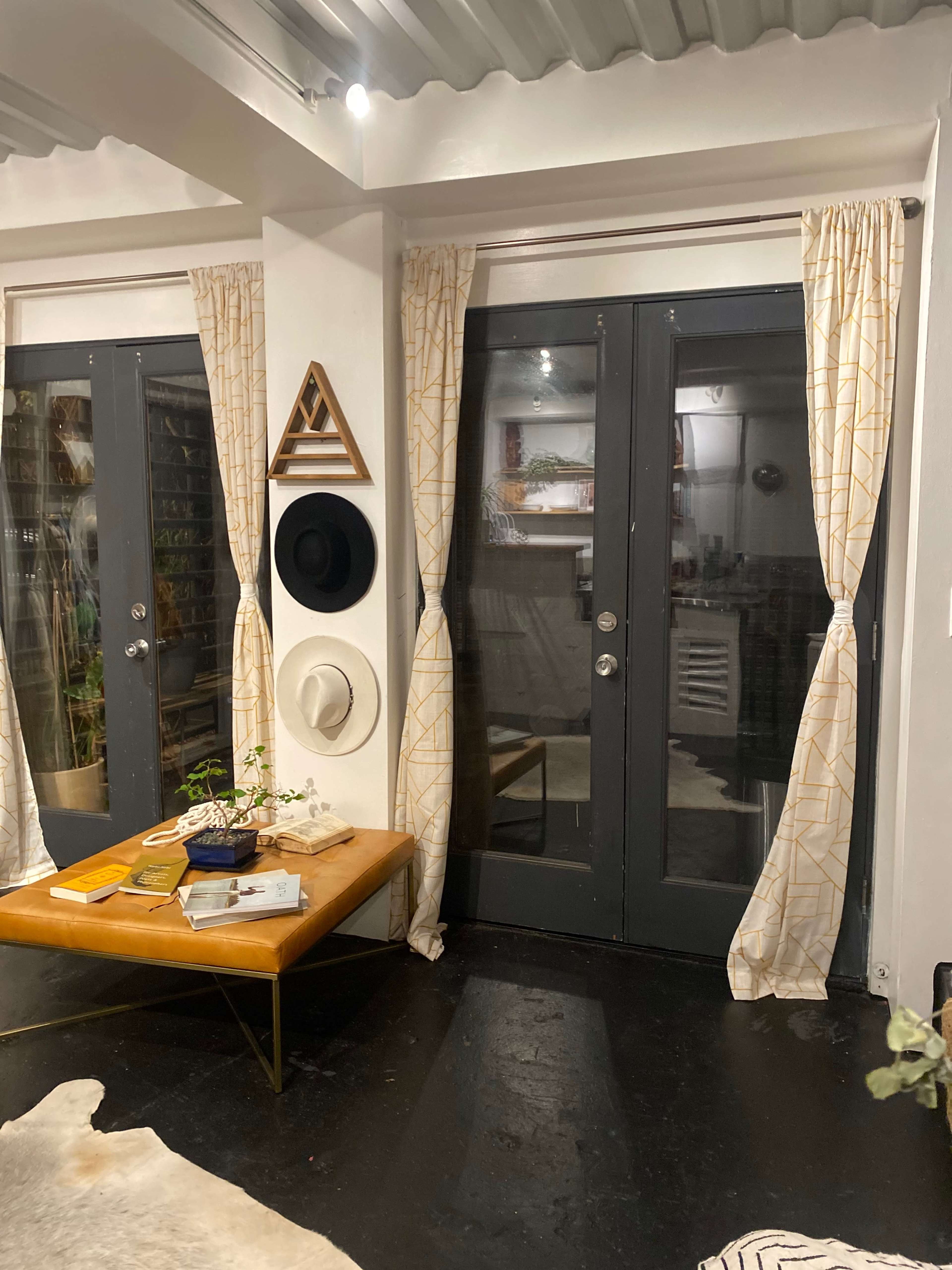 The room features two dark doors with curtains, a coffee table with books, and wall decor including hats and geometric art.