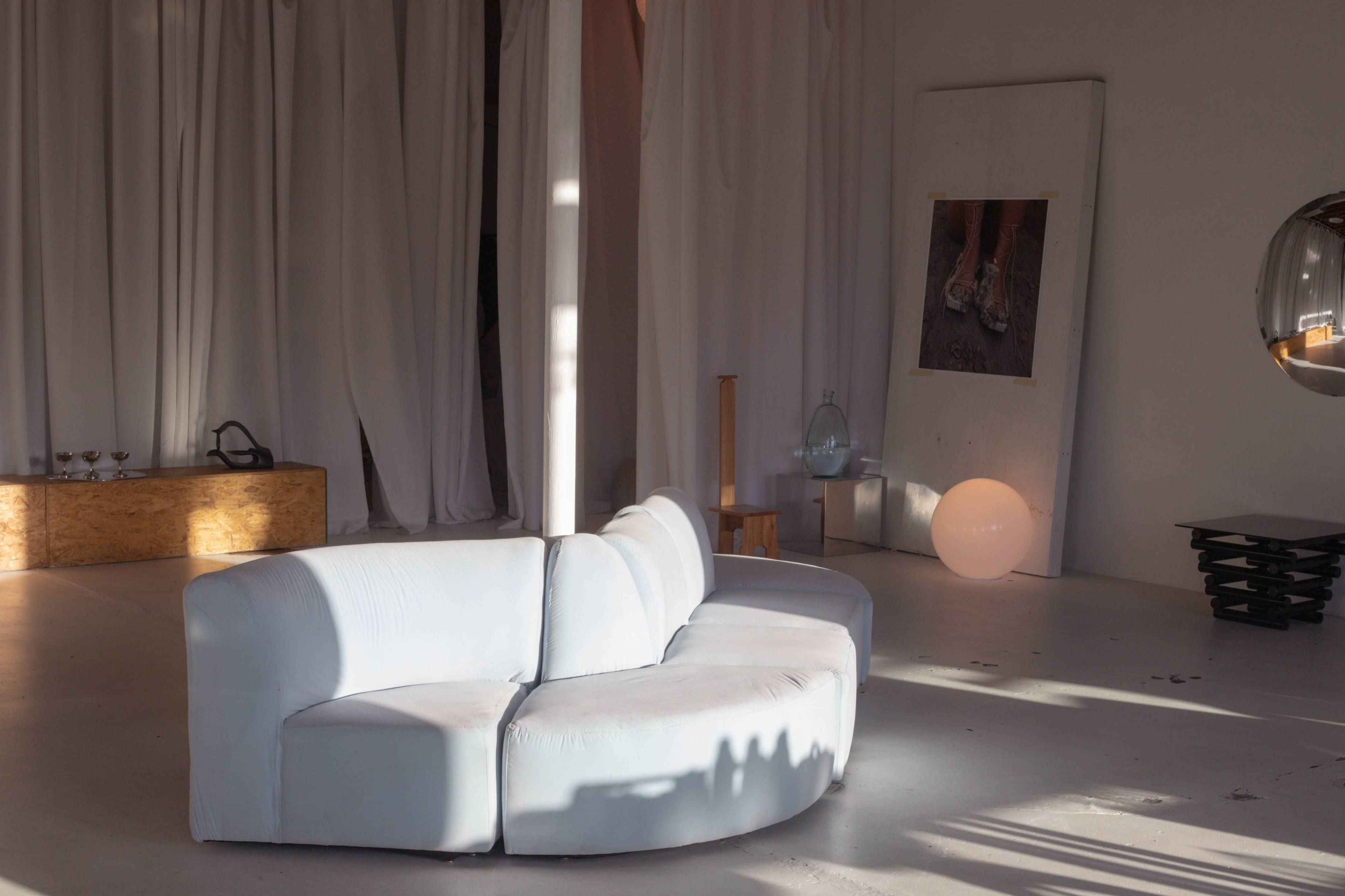A curved, light-colored sofa is positioned in a spacious room with white drapes, a wooden side table, and a large, illuminated orb.