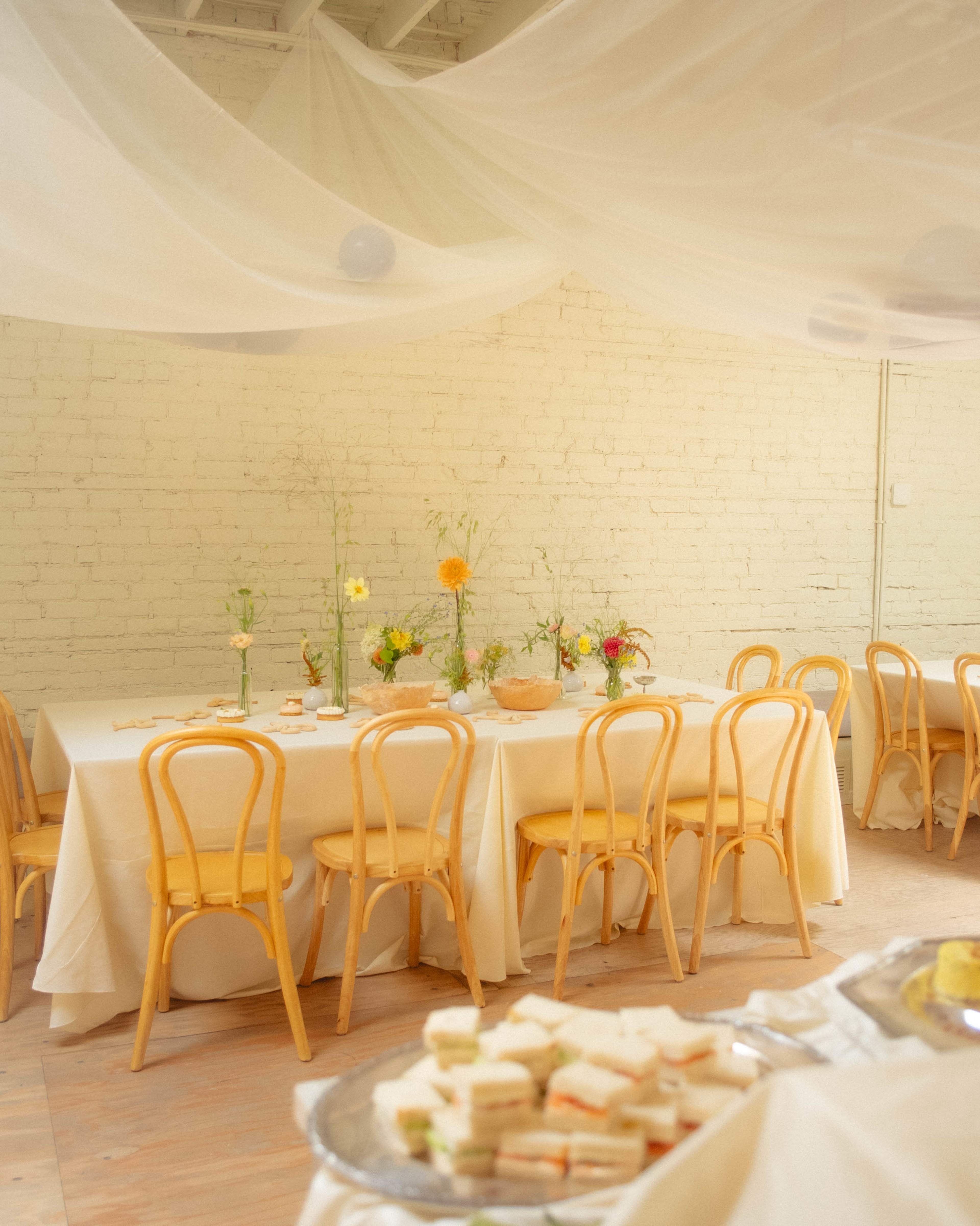 A bright, minimalist dining area features wooden chairs, tablecloths, and floral centerpieces, with a tray of sandwiches visible in the foreground.
