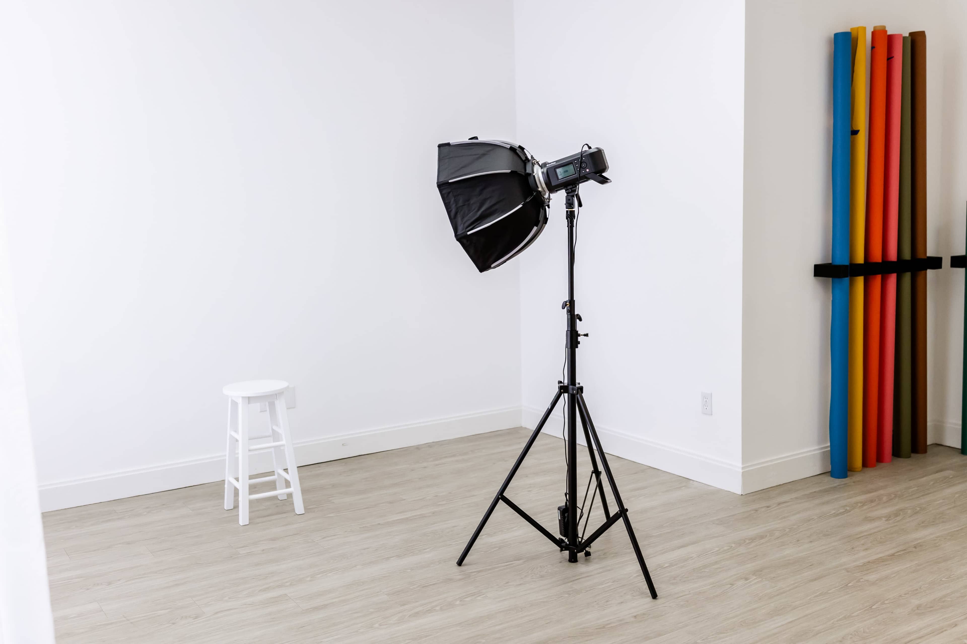 The image shows a photography studio with a light setup positioned next to a white stool and colorful backdrop rolls against the wall.