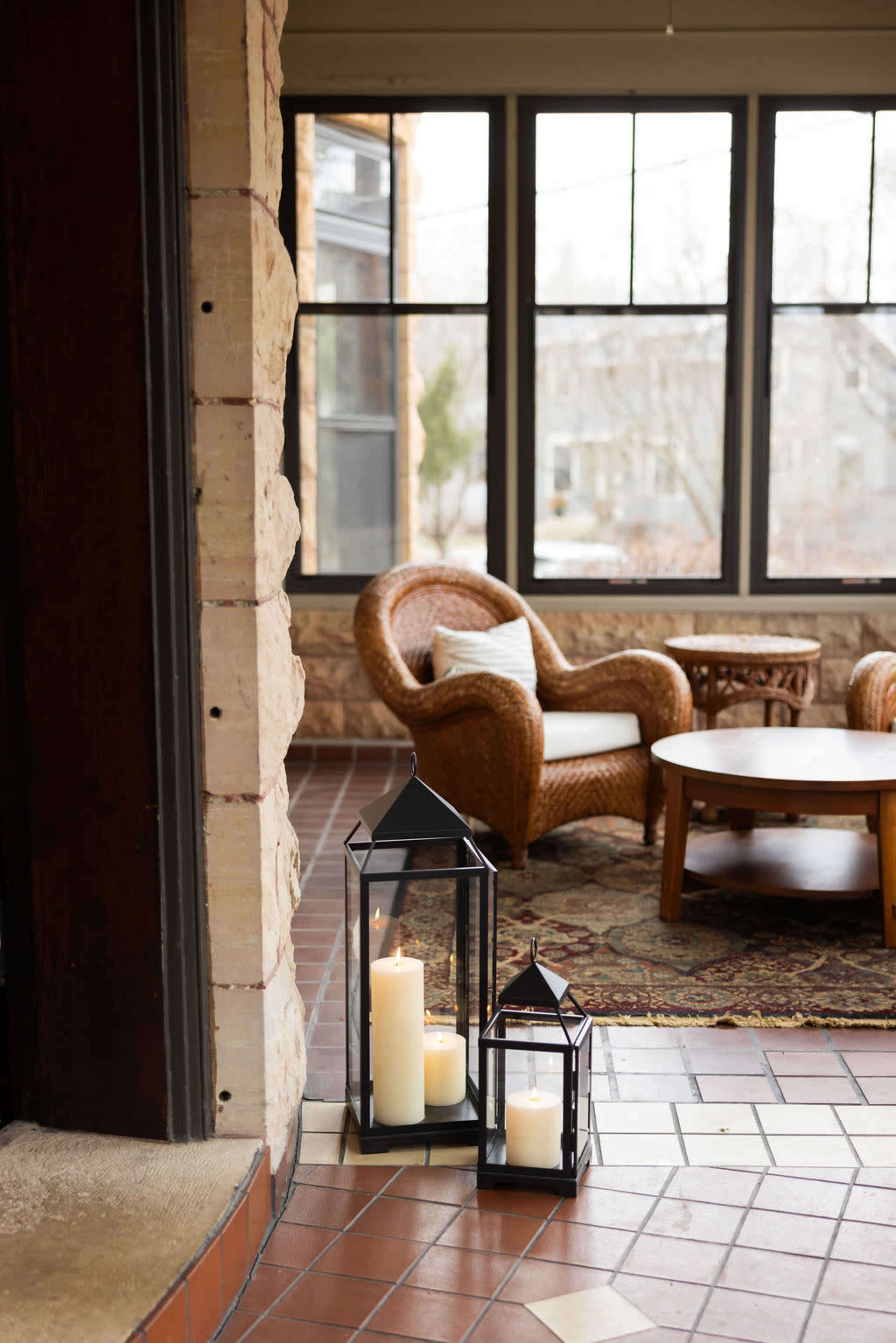 The image shows two lanterns with candles beside a wicker chair and a coffee table in a room with large windows.