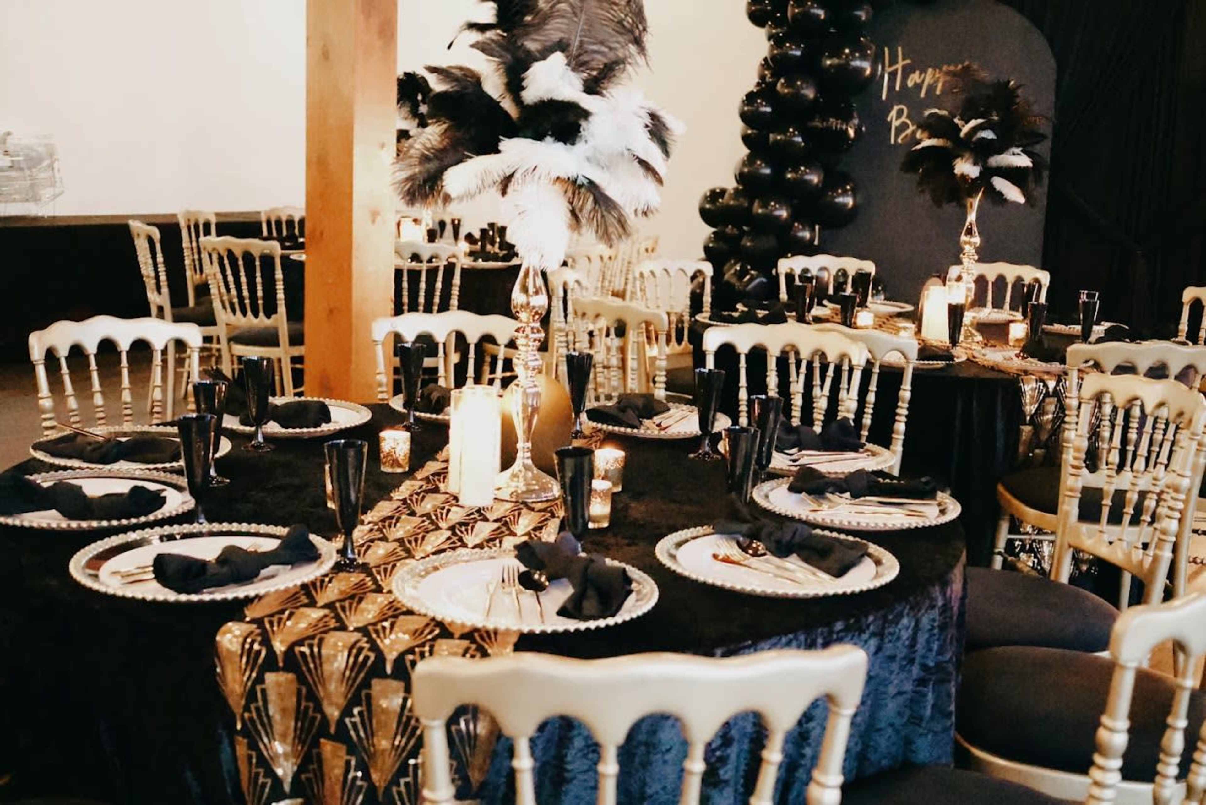 The image shows a elegantly decorated dining area with several tables covered in black fabric, adorned with centerpieces featuring feathers, candles, and ornate table settings.