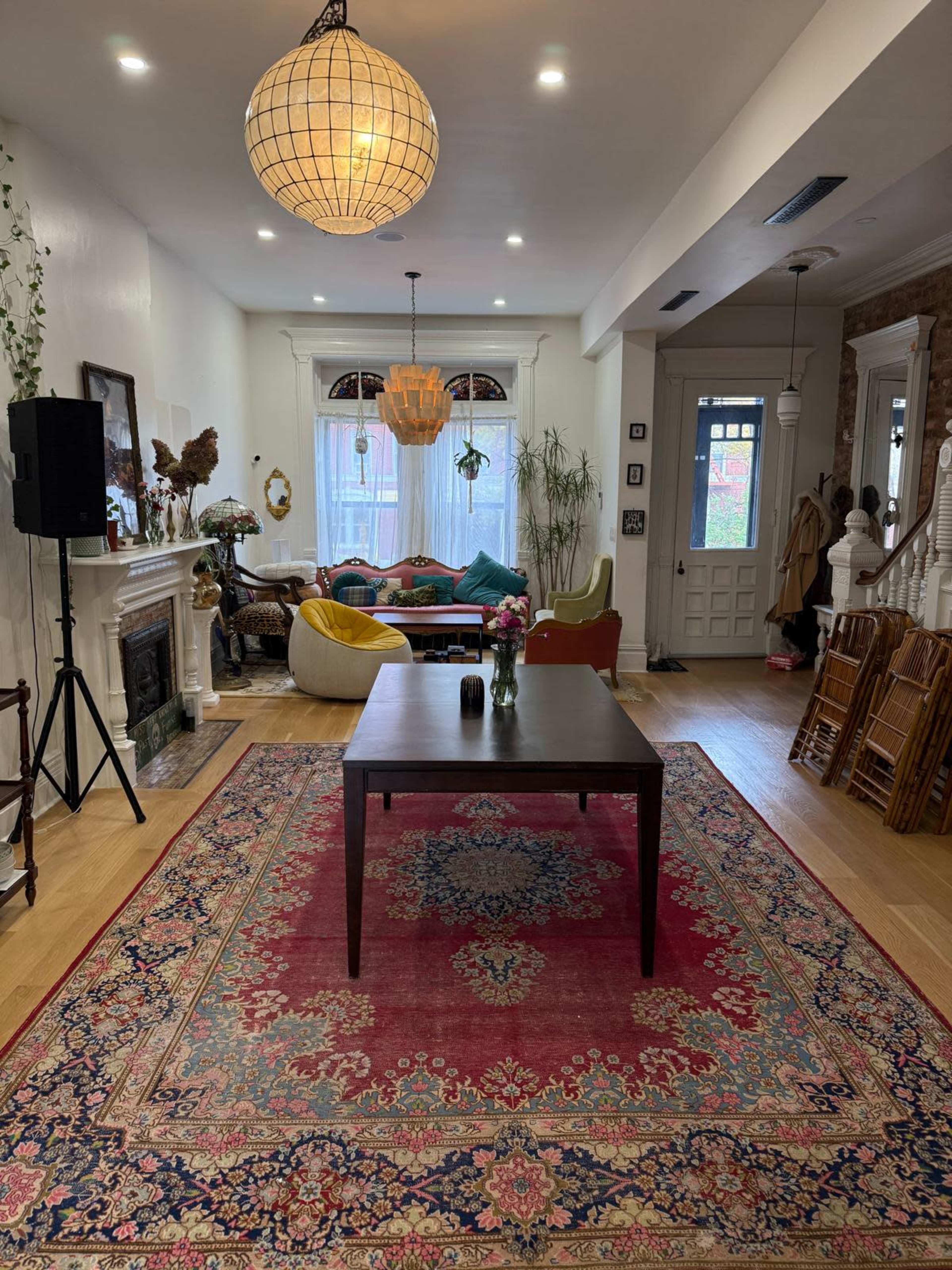 The image shows a spacious living area with a large, ornate rug, a central table, a chandelier, and a mix of colorful seating arrangements.
