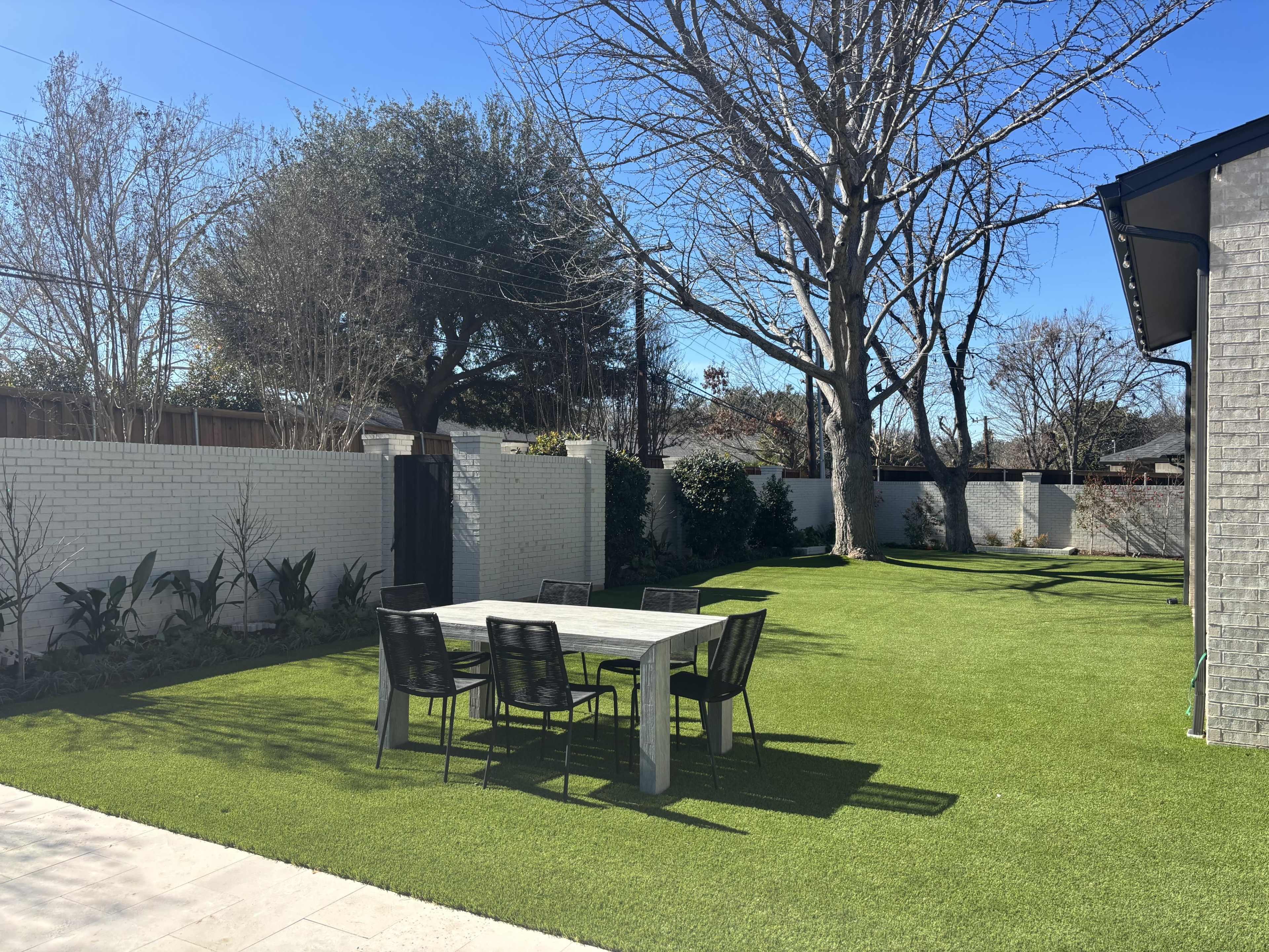 The image shows a backyard with artificial grass, a concrete dining table, and several black chairs surrounded by trees and shrubs.