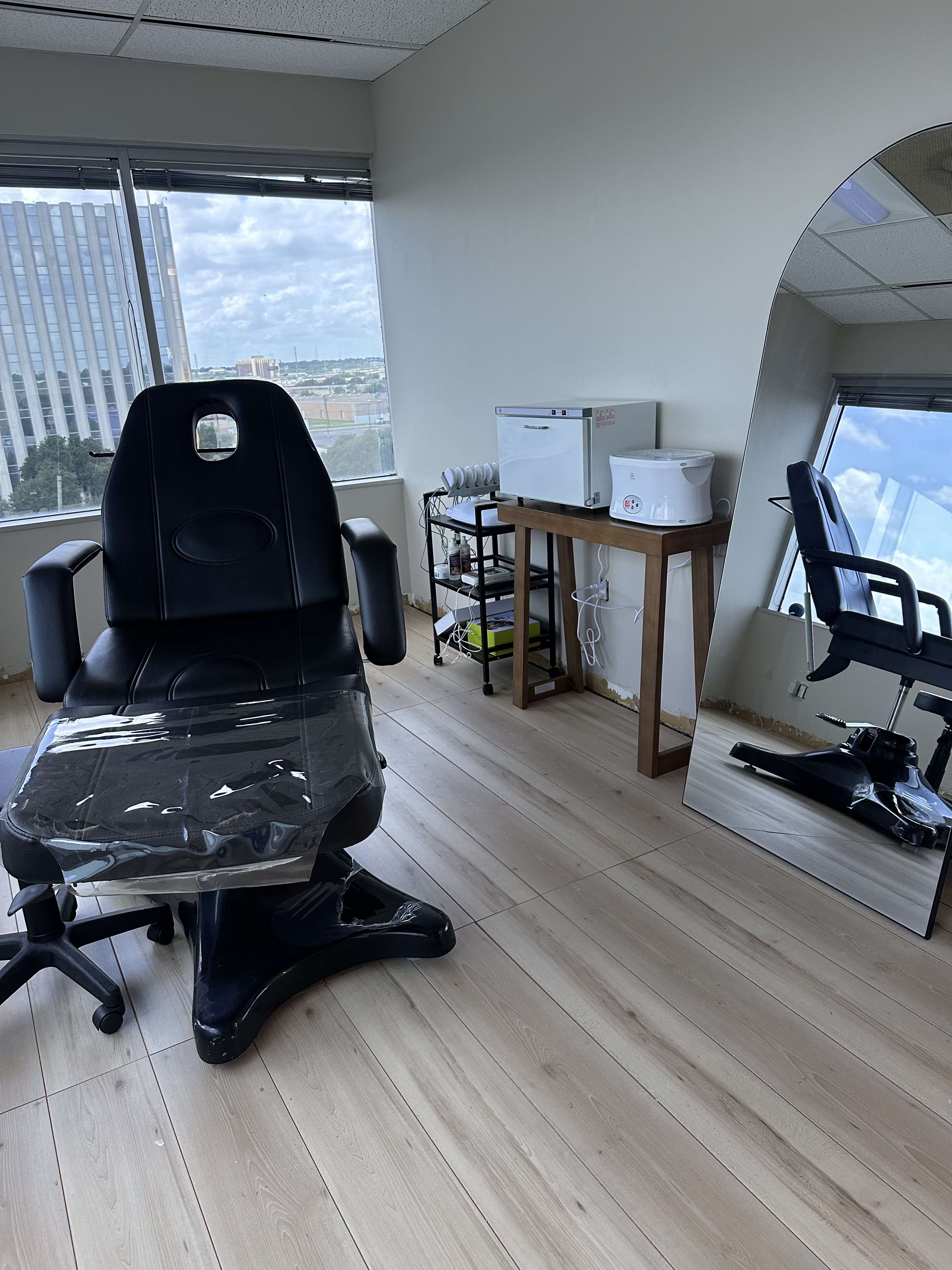 The image shows a professional salon or spa room with a black treatment chair, a mirror, and a wooden table equipped with various tools and supplies.