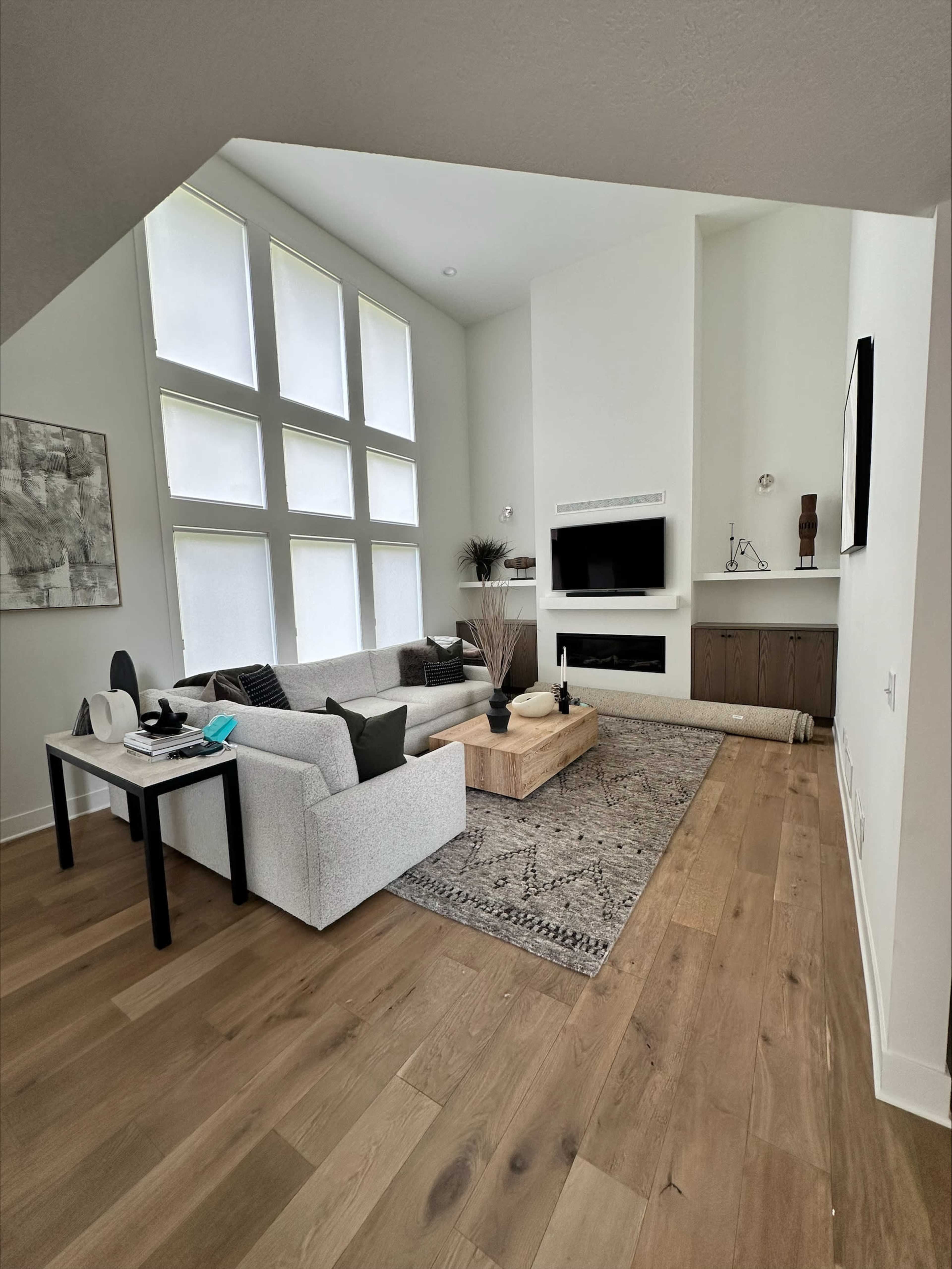 A modern living room featuring a light gray sectional sofa, a wooden coffee table, a large area rug, and large windows that allow natural light to enter the space.