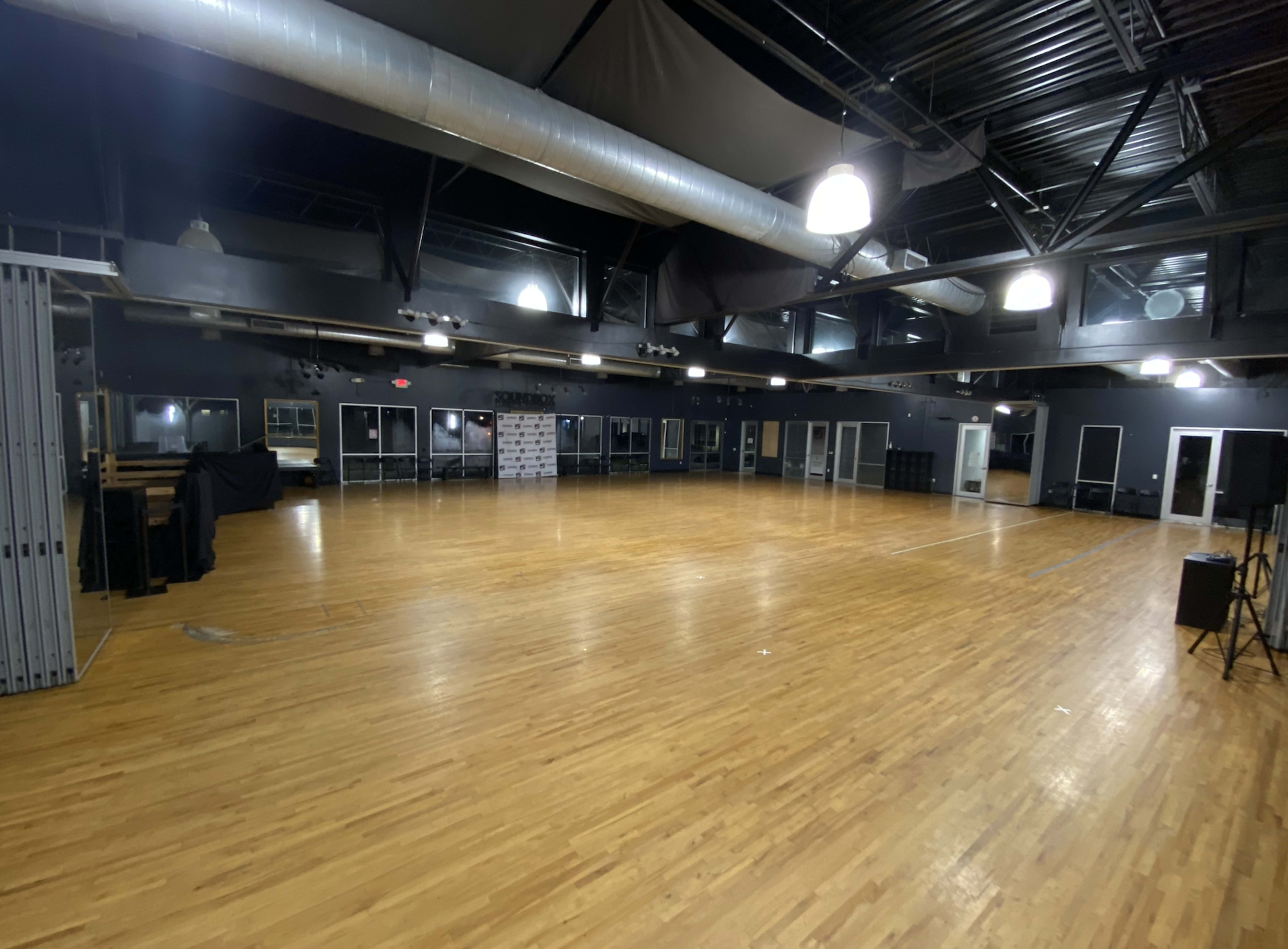 An empty, spacious indoor venue with a wooden floor and a black ceiling, featuring large windows and stage lighting.