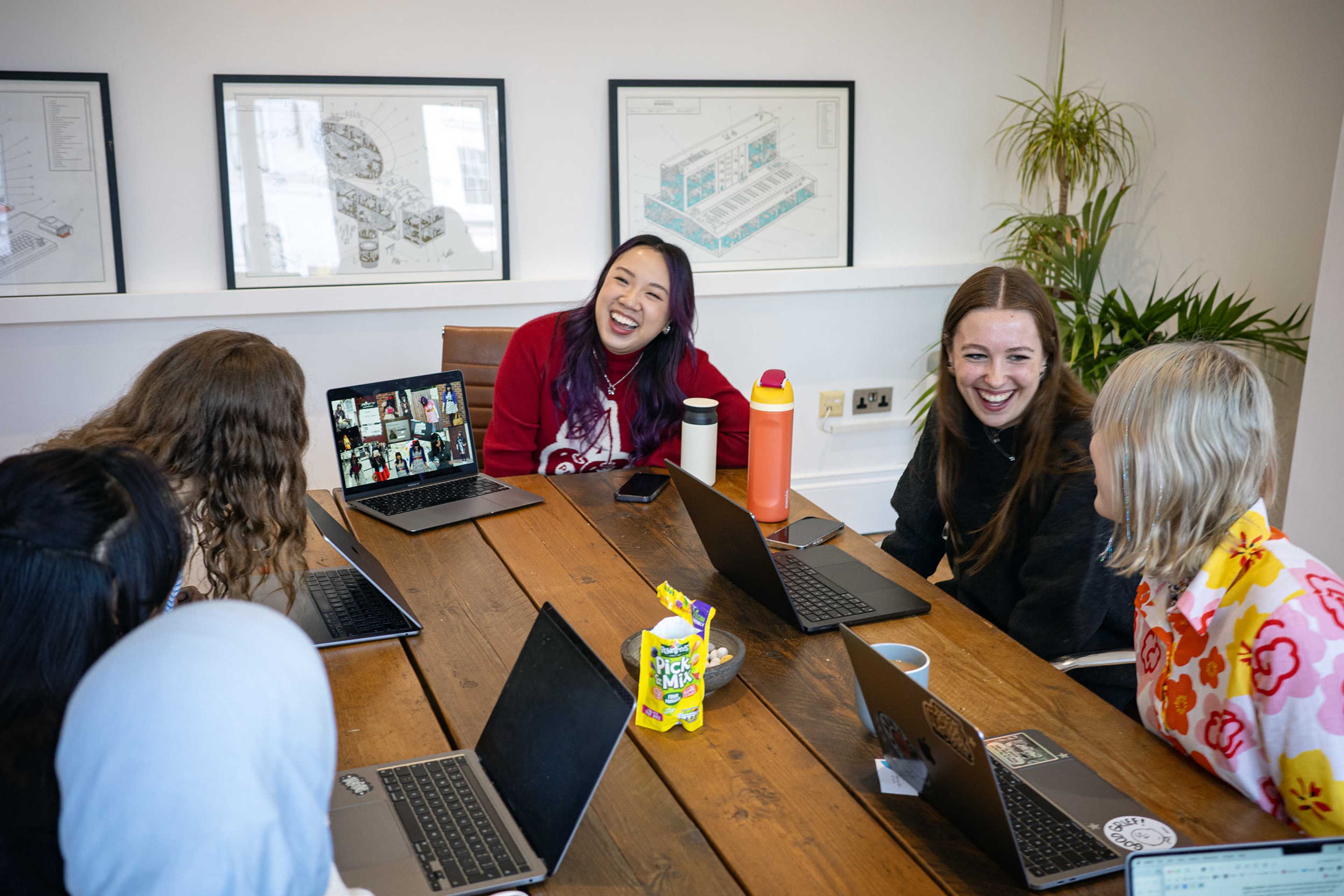A group of seven people sits around a wooden table in a bright room, engaging with each other while using laptops.