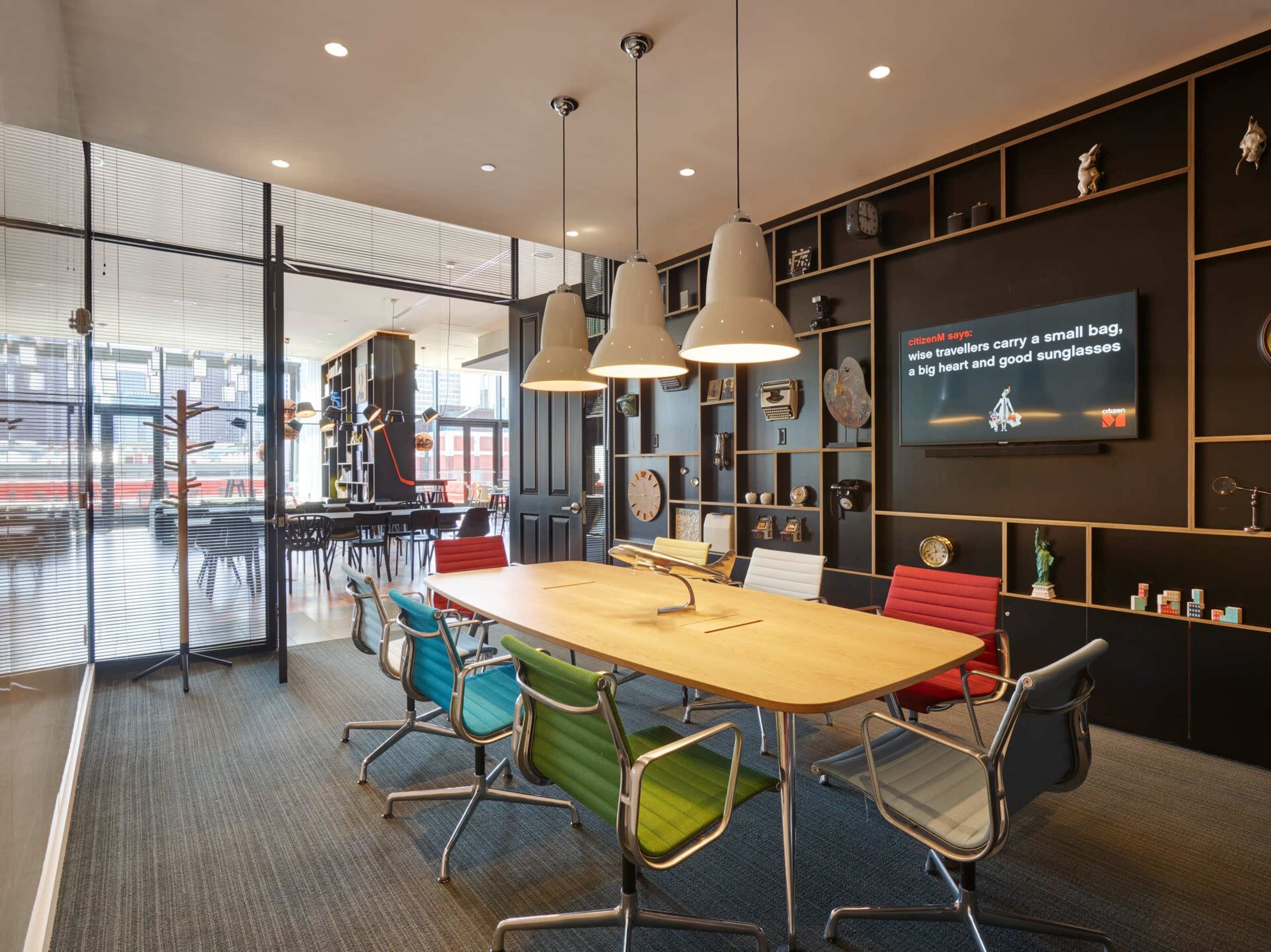 A modern conference room features a large wooden table surrounded by colorful chairs and a wall-mounted screen, with floor-to-ceiling windows providing a view of the outside.