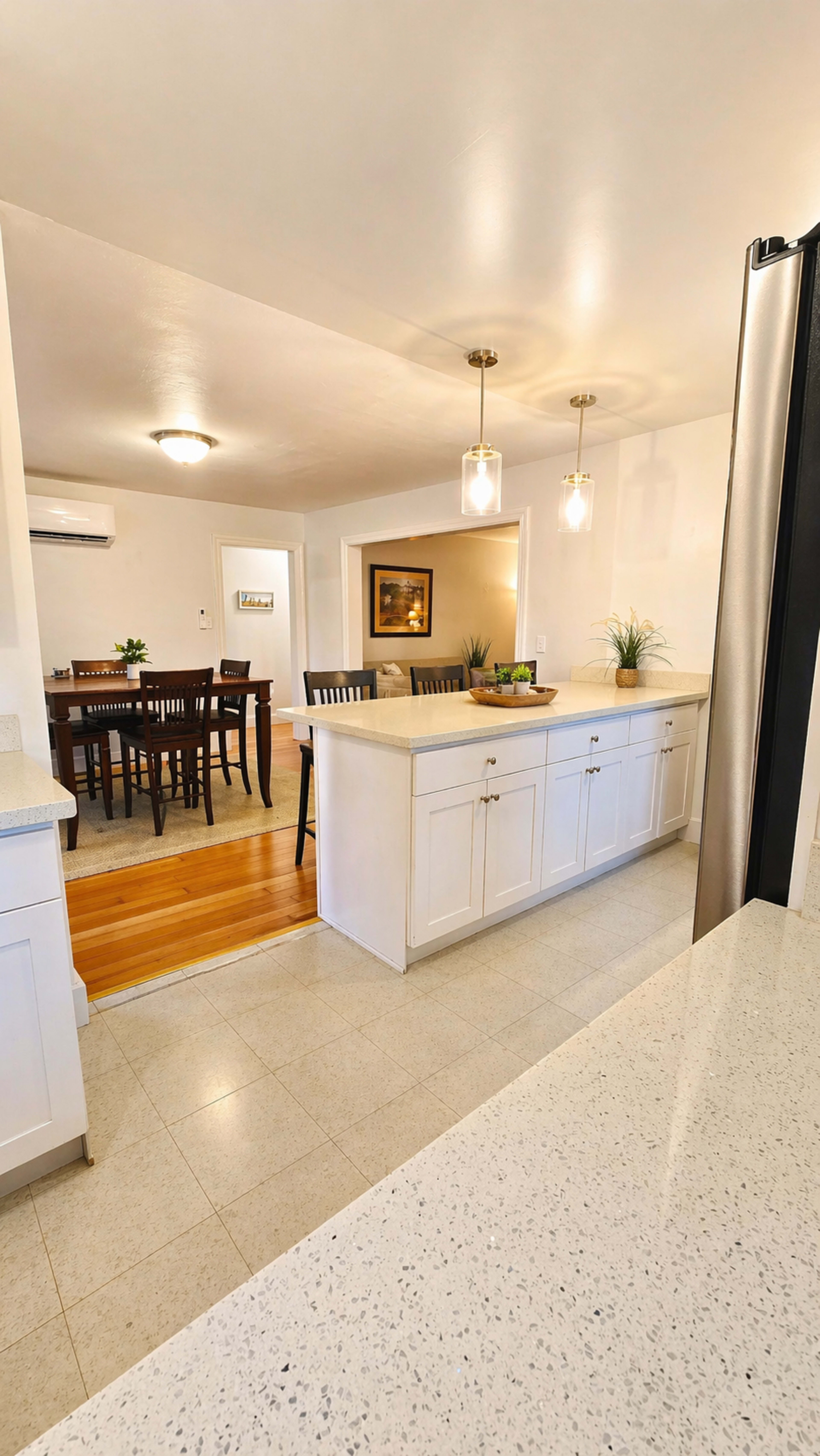 The image shows a modern kitchen with white cabinets and a countertop, leading to a dining area with a wooden table and chairs.