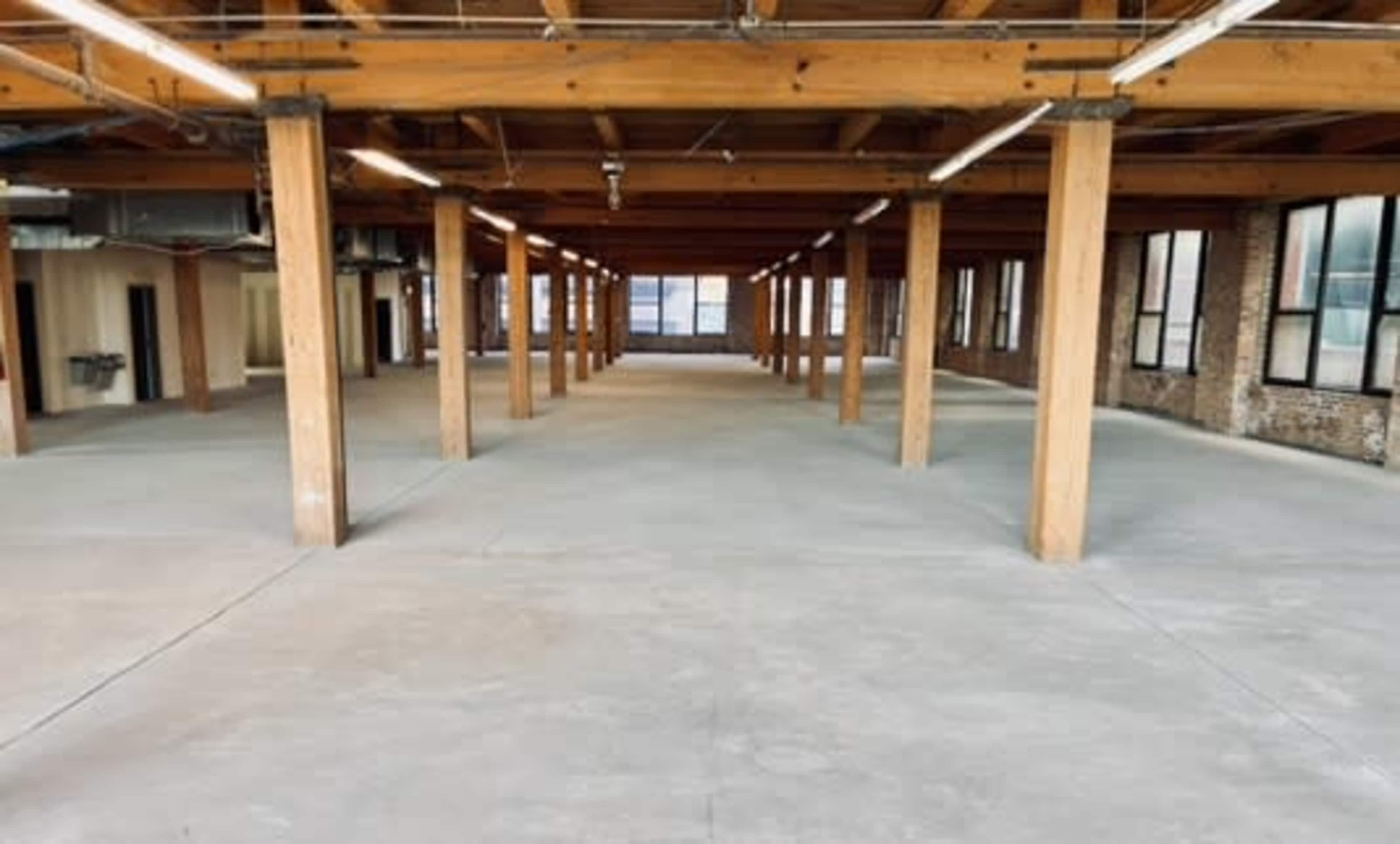The image shows a spacious, empty interior of a building with wooden beams and concrete flooring.