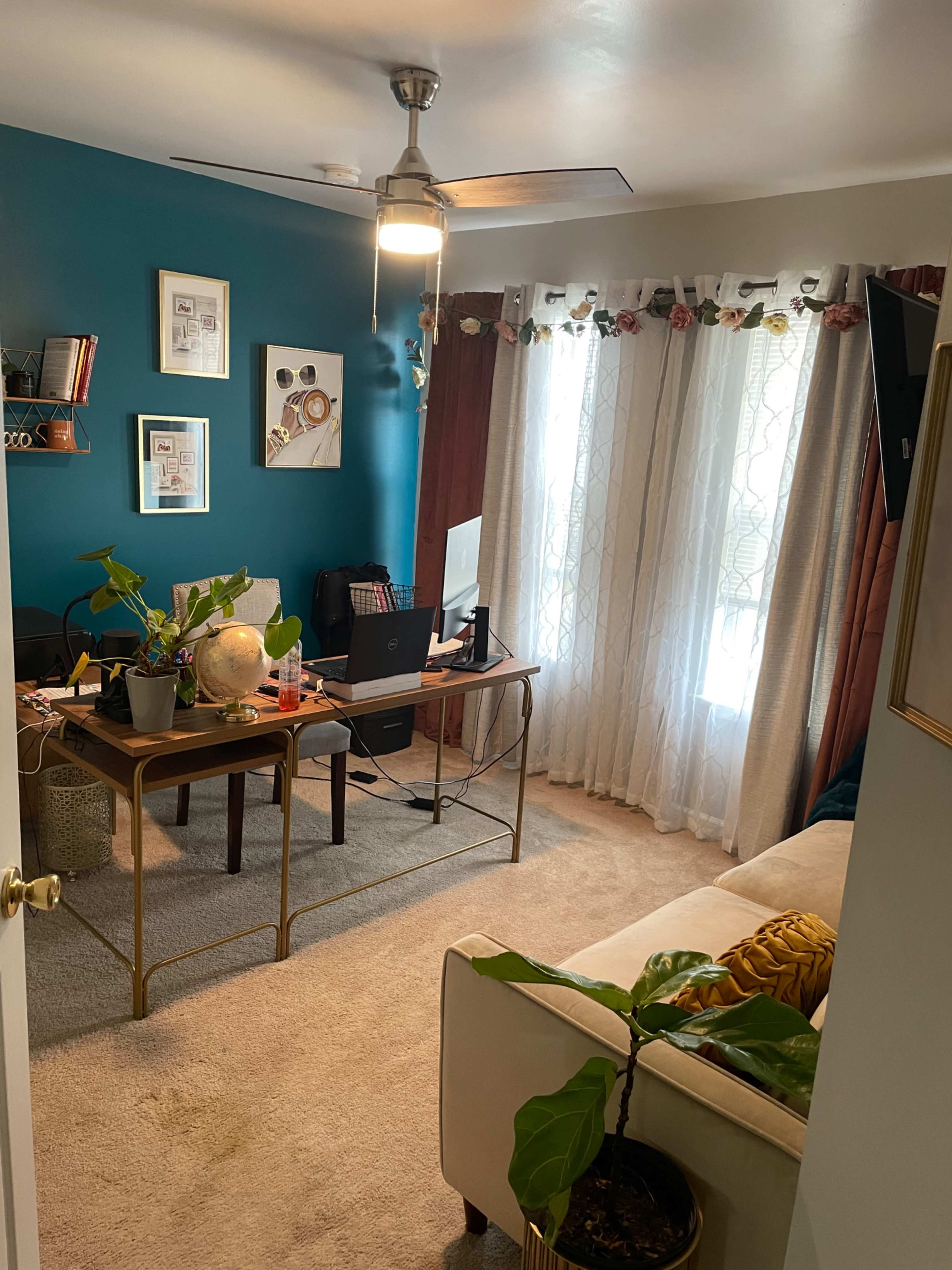 A cozy home office with a desk, computer, and various plants, set against a teal wall and bright window treatments.
