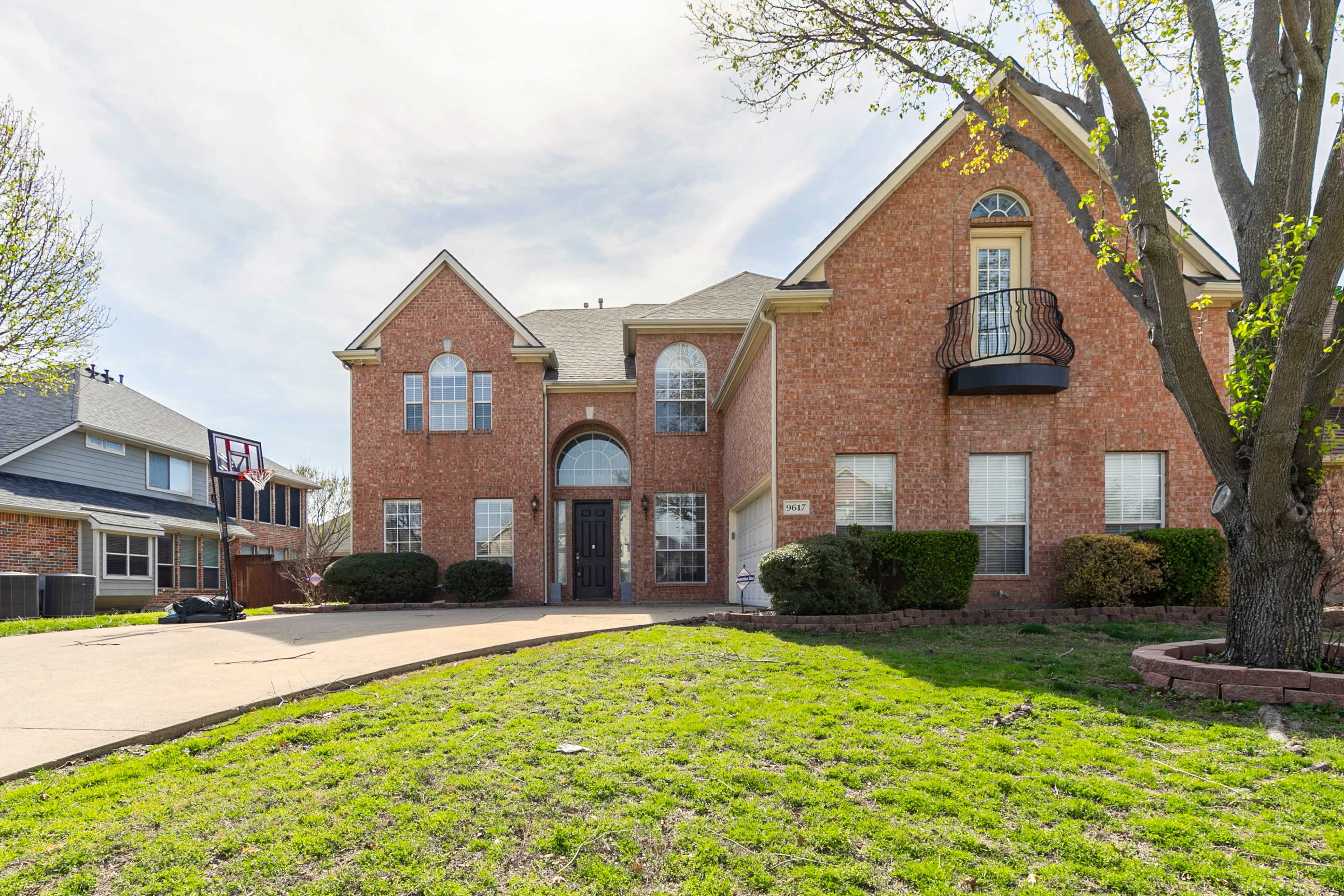 A two-story brick house with a front balcony, a well-manicured lawn, and a driveway leading to the entrance.