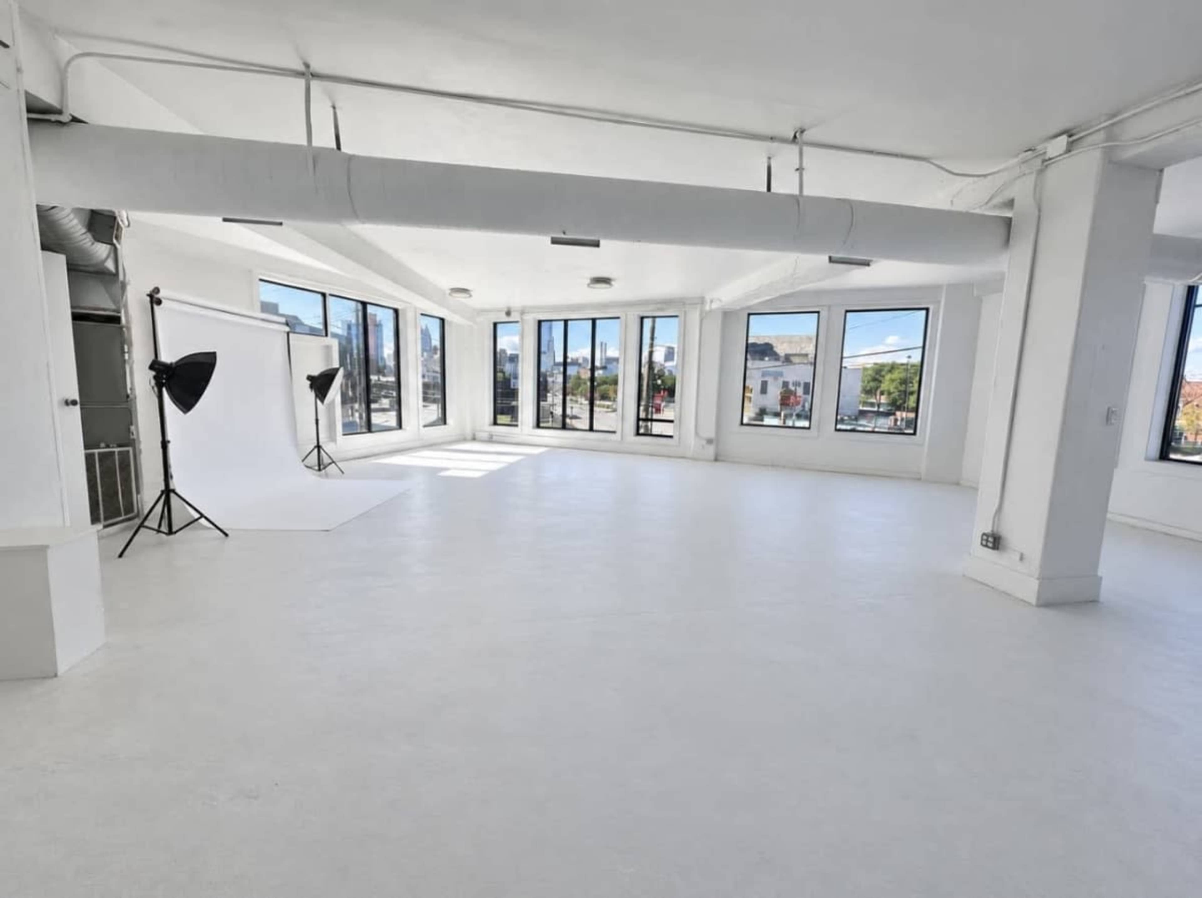 The image shows a spacious, well-lit studio with large windows, white walls, and a minimalistic setup including a backdrop and lighting equipment.