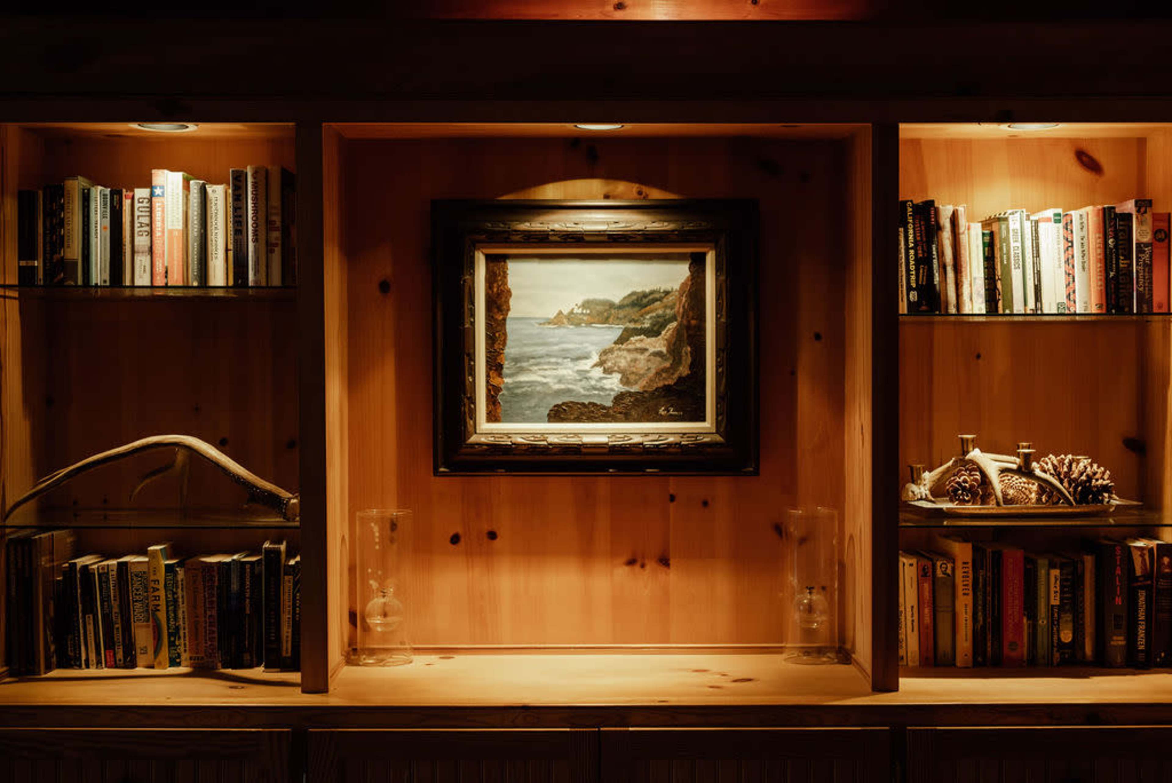 The image shows a wooden bookshelf with books on either side and a framed painting of a coastal scene centered above.