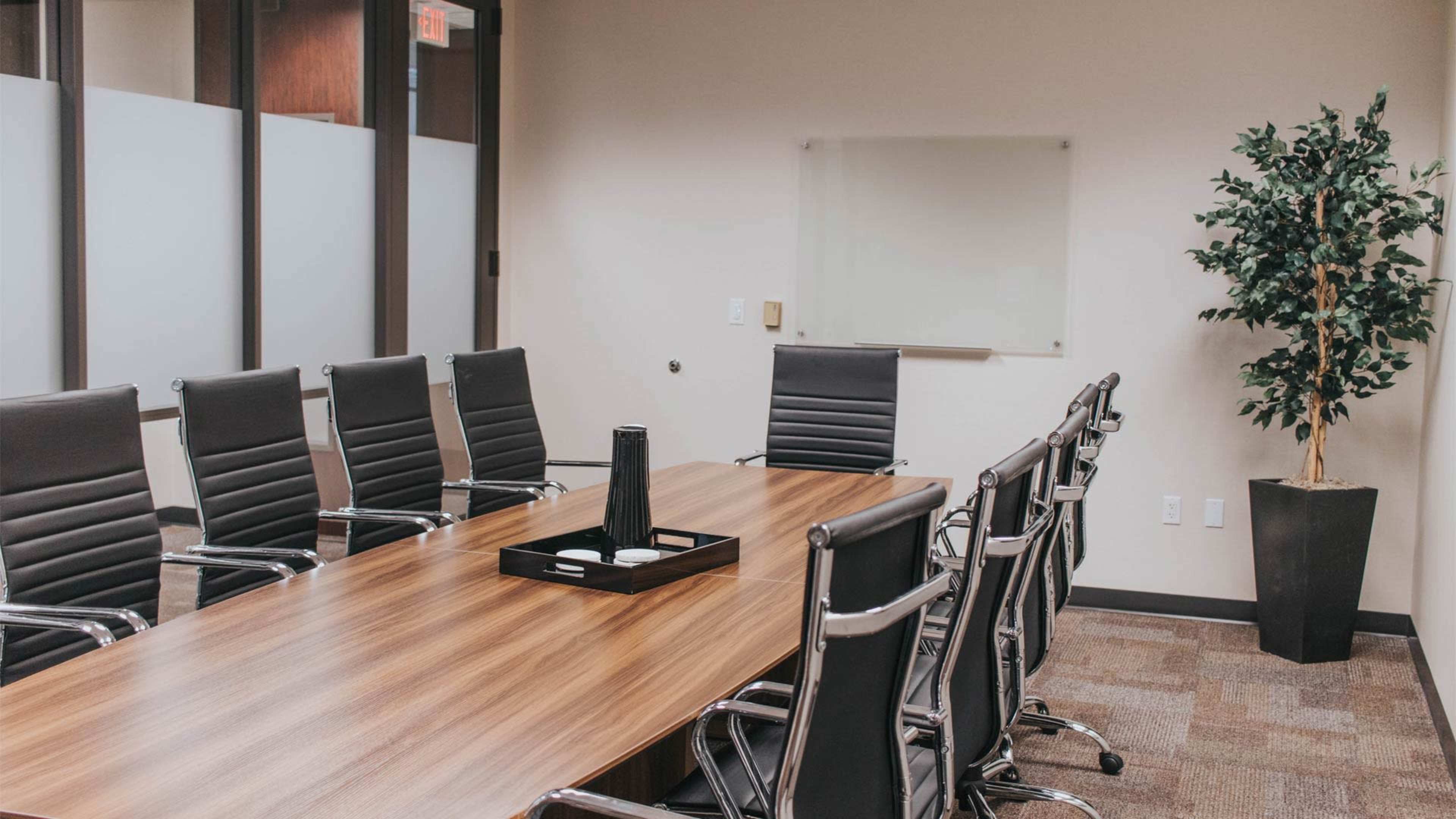 A modern conference room features a long wooden table surrounded by black ergonomic chairs, with a potted plant in one corner and a glass whiteboard on the wall.