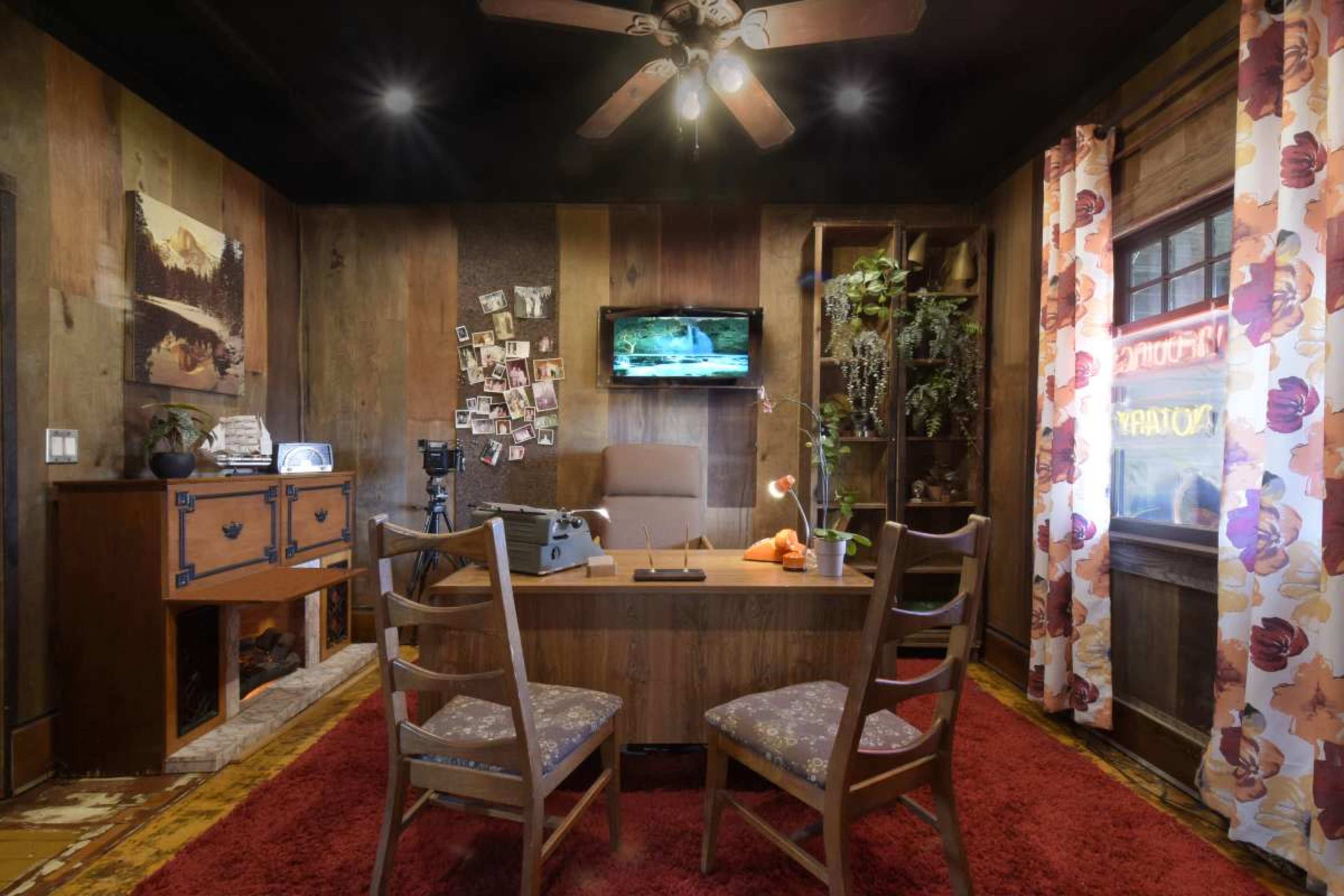 The image depicts a cozy office space featuring a wooden desk, two chairs, and a mounted television, surrounded by wood-paneled walls and decorated with plants and photographs.