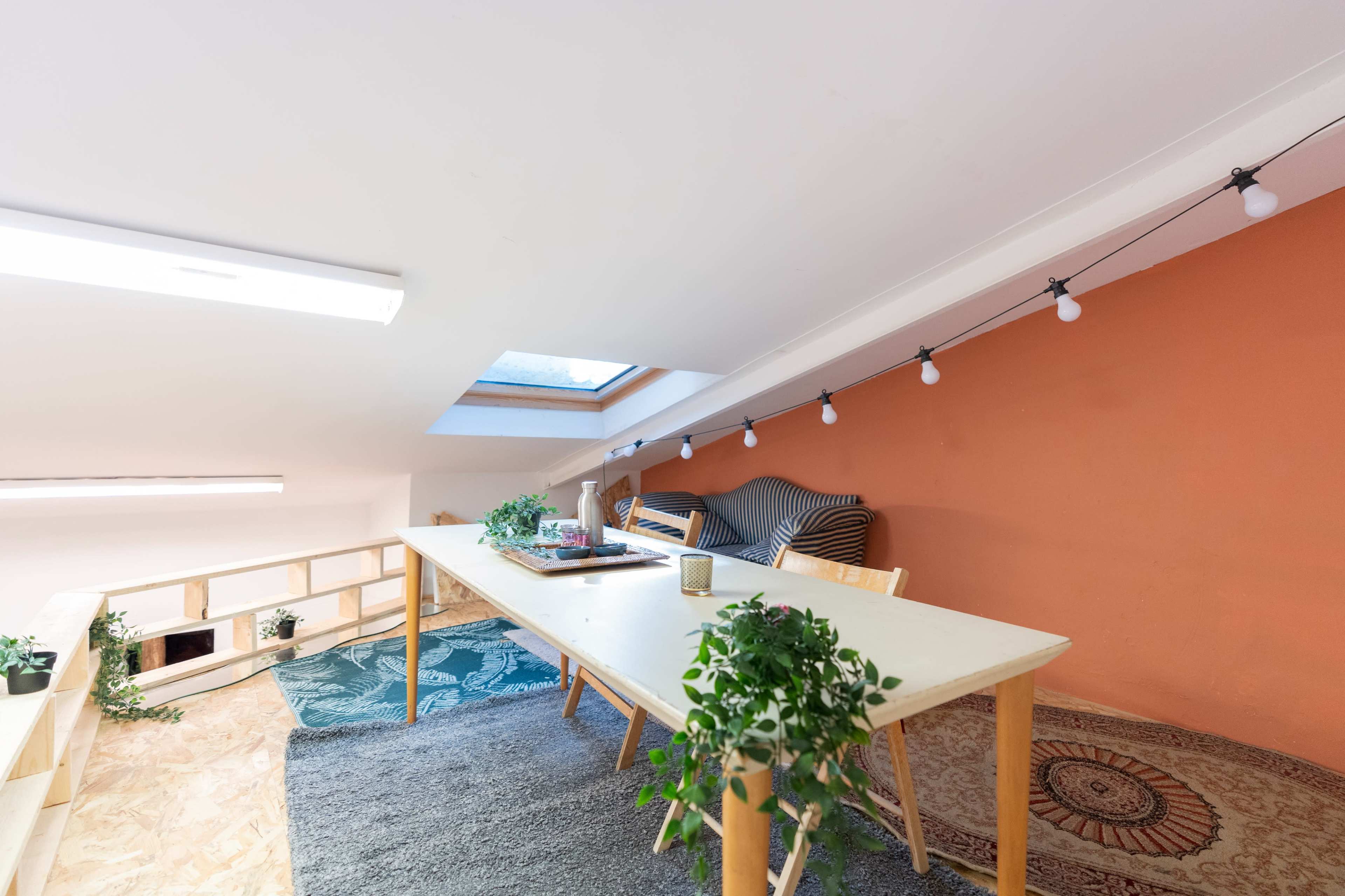 An attic space features a long table surrounded by chairs, with a cozy seating area and decorative plants under a skylight.