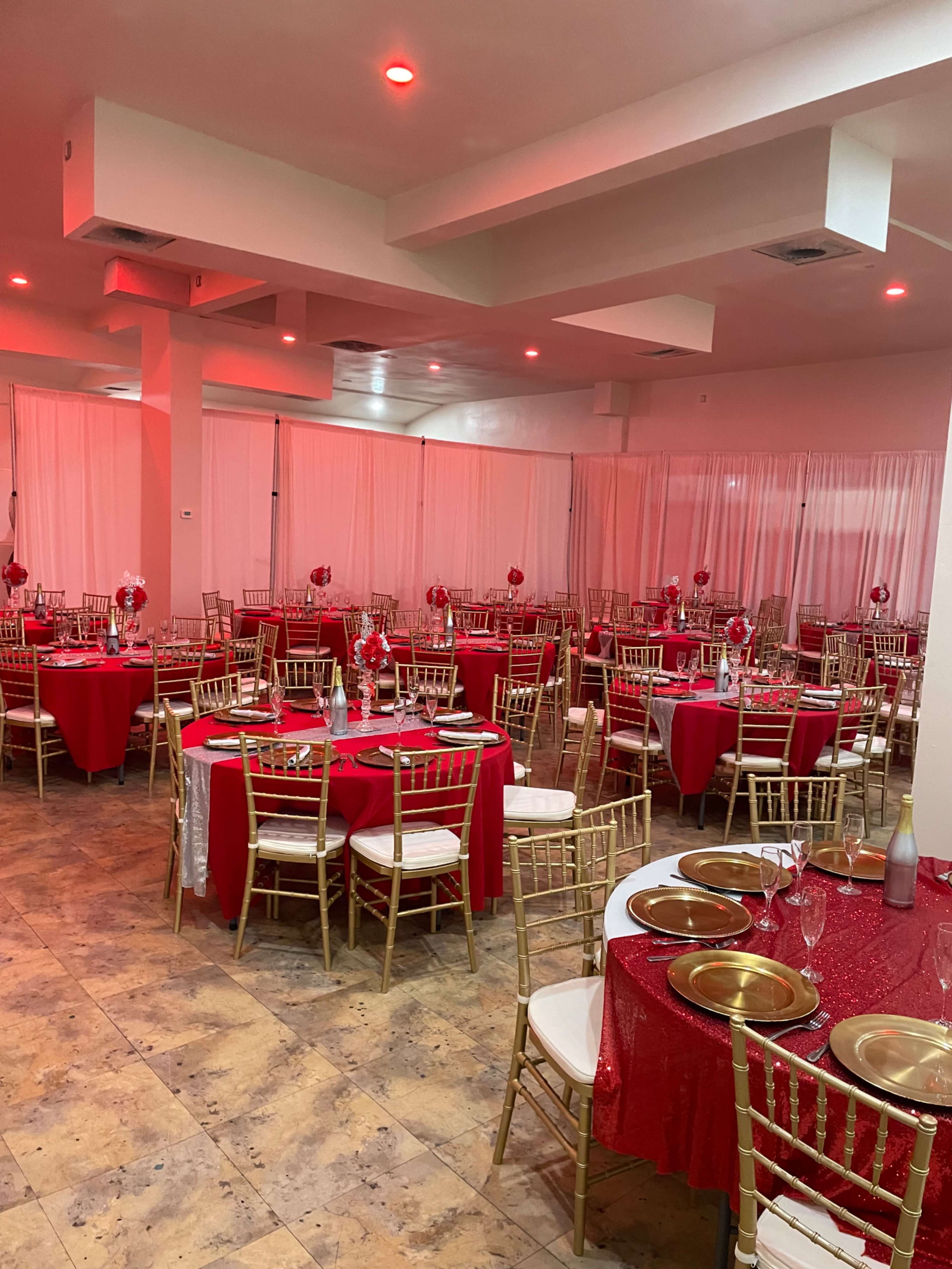 The image shows an elegantly decorated dining area with round tables covered in red tablecloths, gold accents, and surrounding gold chiavari chairs, illuminated by soft red lighting.