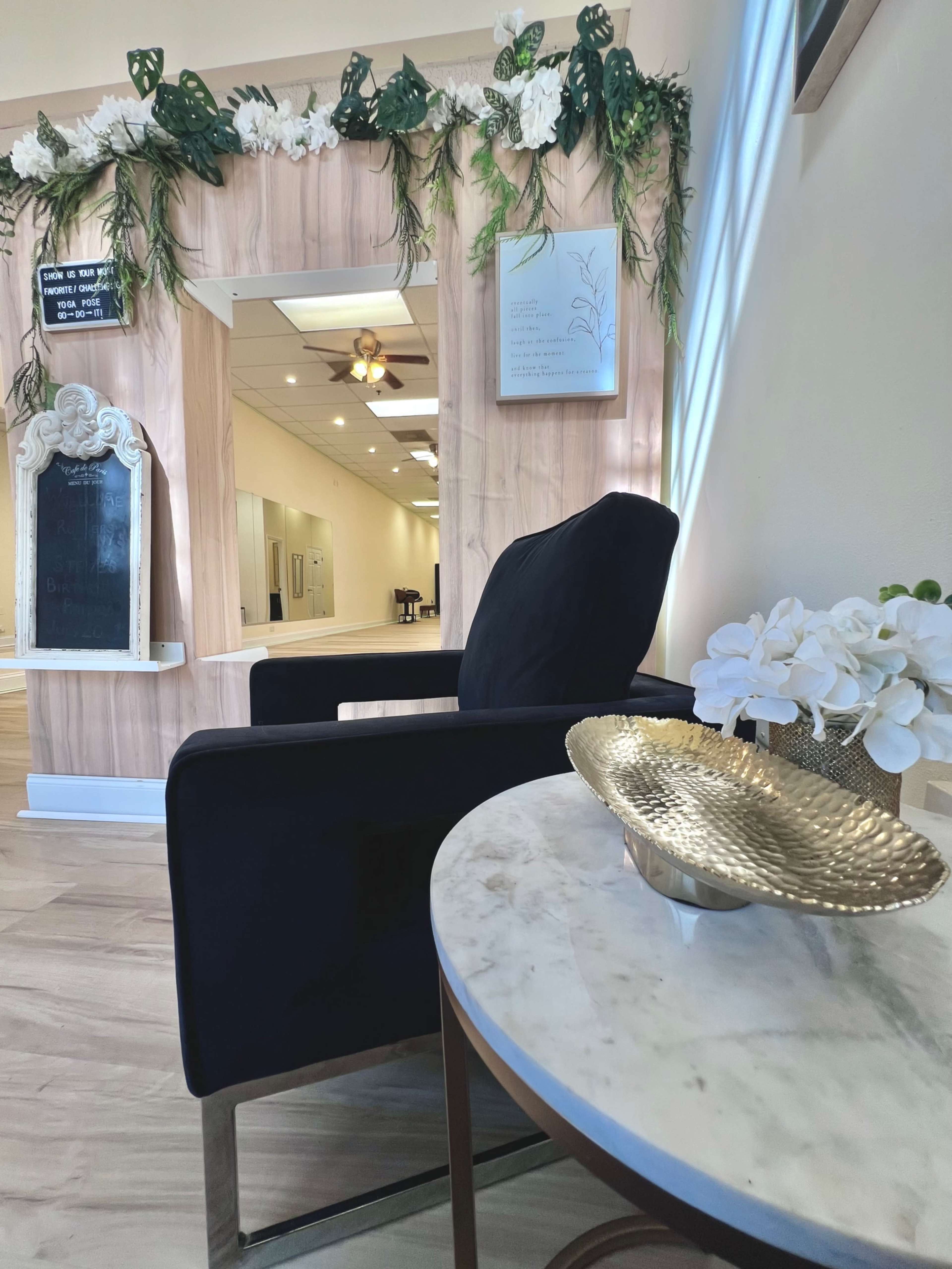 The image shows a modern interior setting featuring a black armchair next to a round table with a decorative gold bowl and a small white flower arrangement, against a backdrop of wood paneling and greenery.