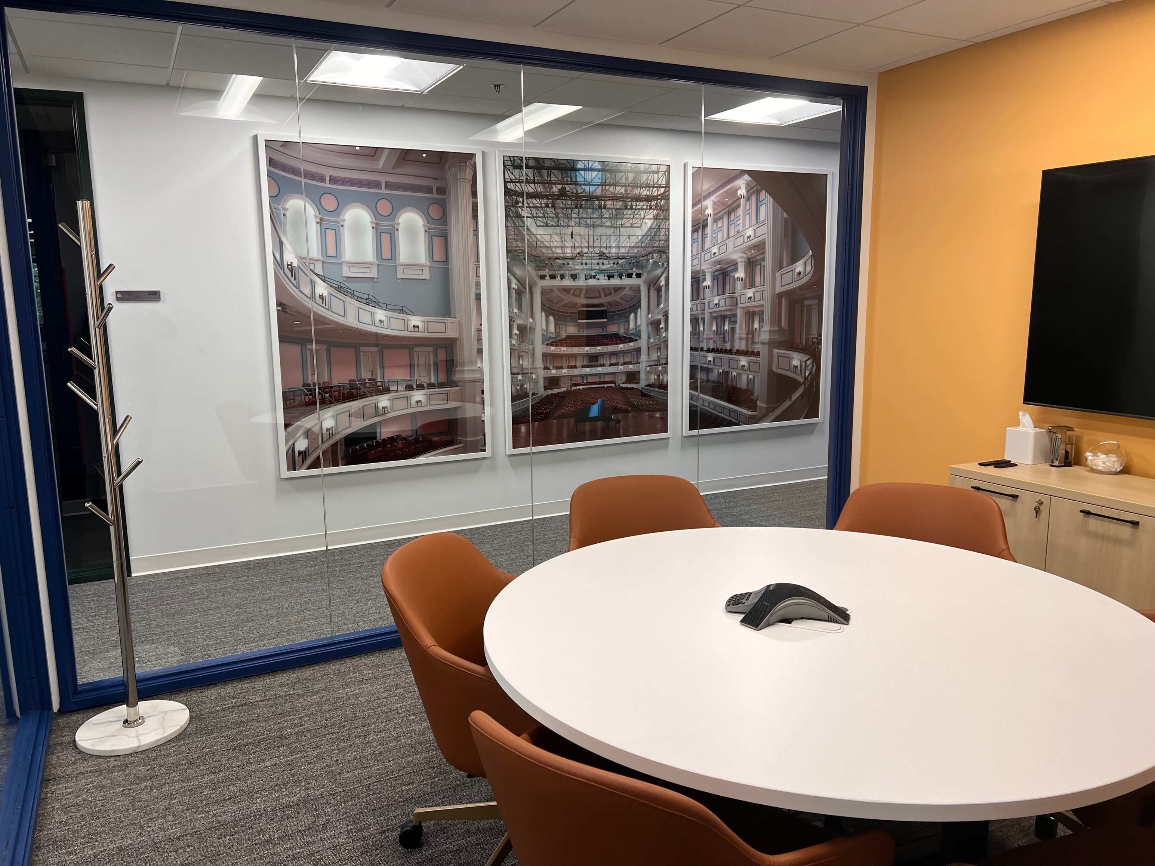 A conference room with a round white table surrounded by orange chairs and large framed photographs of a theater on the walls.