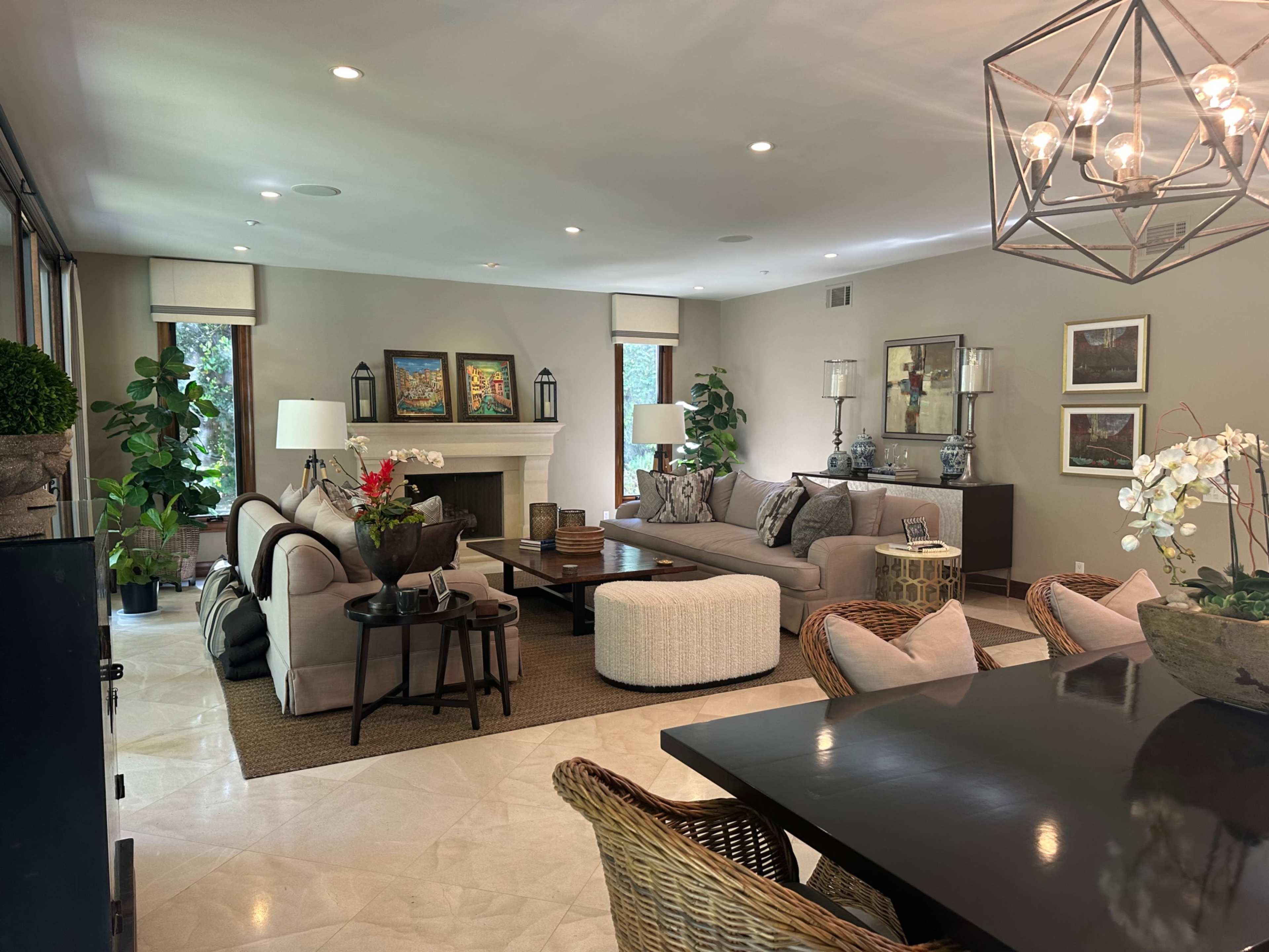 The image depicts a spacious living area featuring a beige sofa, a round pouf, a dining table, and large windows with plants, framed artwork, and decorative lamps.