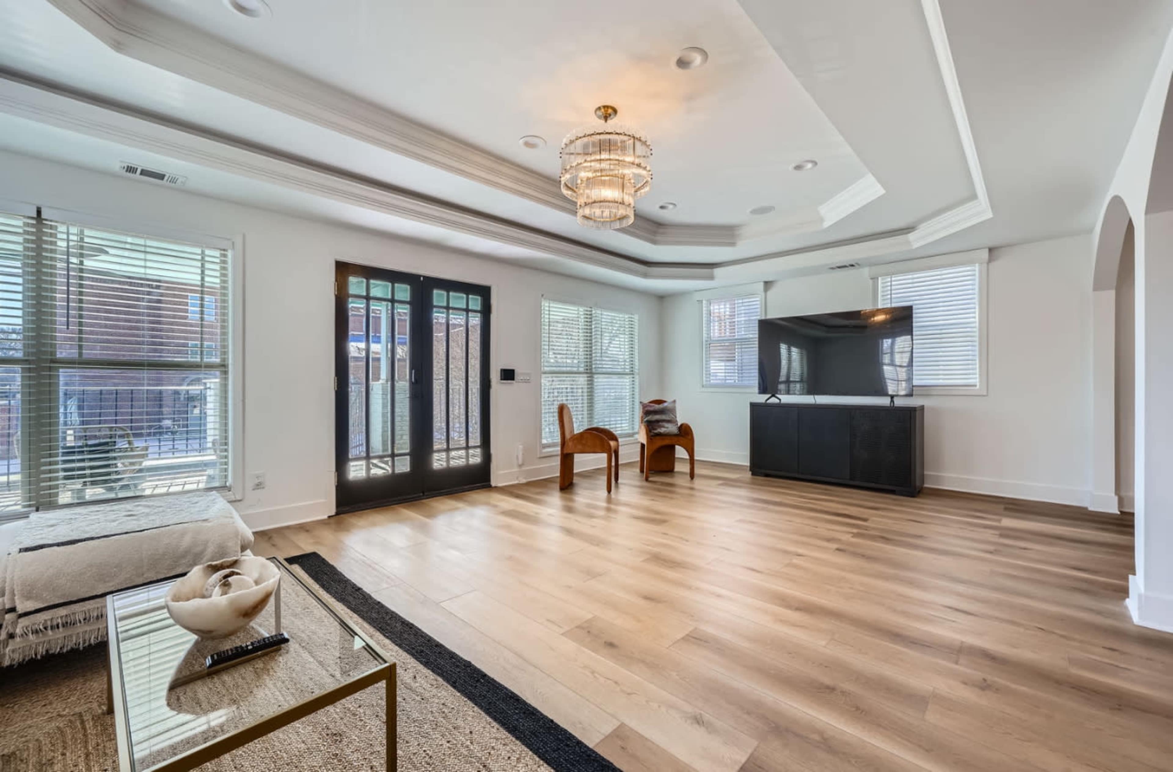 The image shows a modern living room with wooden flooring, large windows, and a central chandelier, featuring a pair of chairs and a television unit.