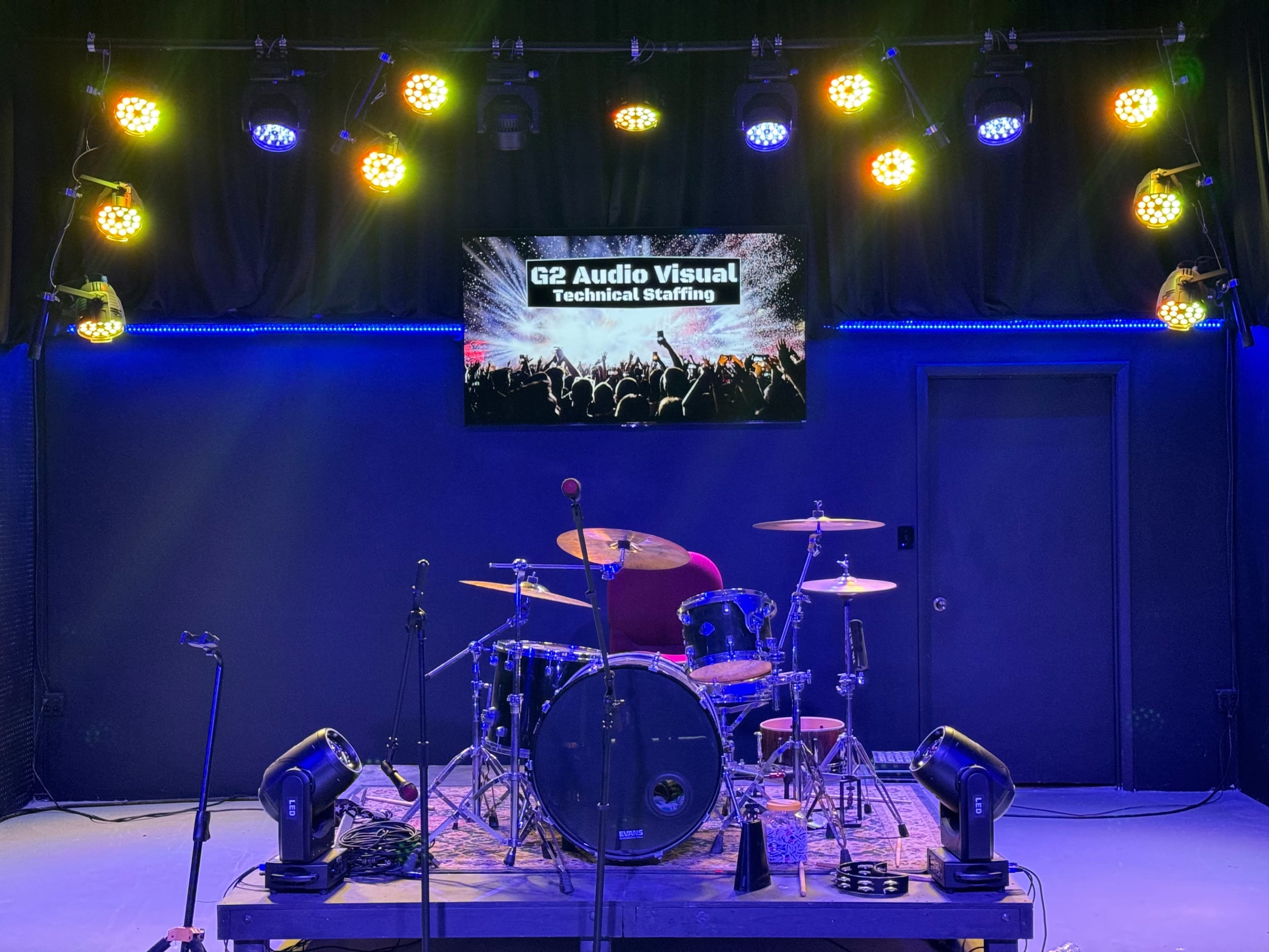 The image shows a stage setup with a drum kit, microphones, and colorful stage lights, while a screen displays "G2 Audio Visual Technical Staffing."