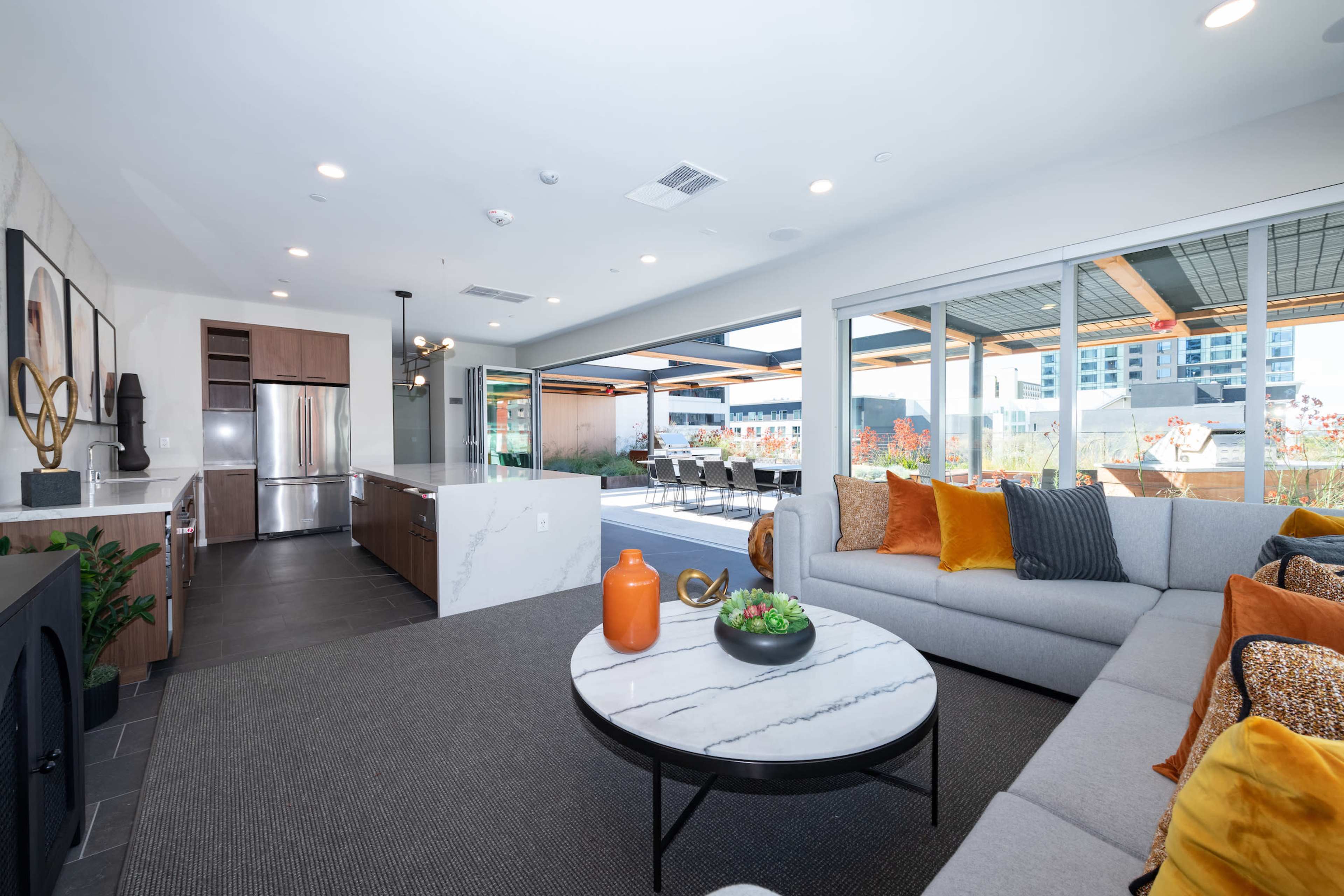 A modern living space with a gray sectional sofa, a marble coffee table, and a kitchen area featuring stainless steel appliances and large windows overlooking an outdoor dining area.