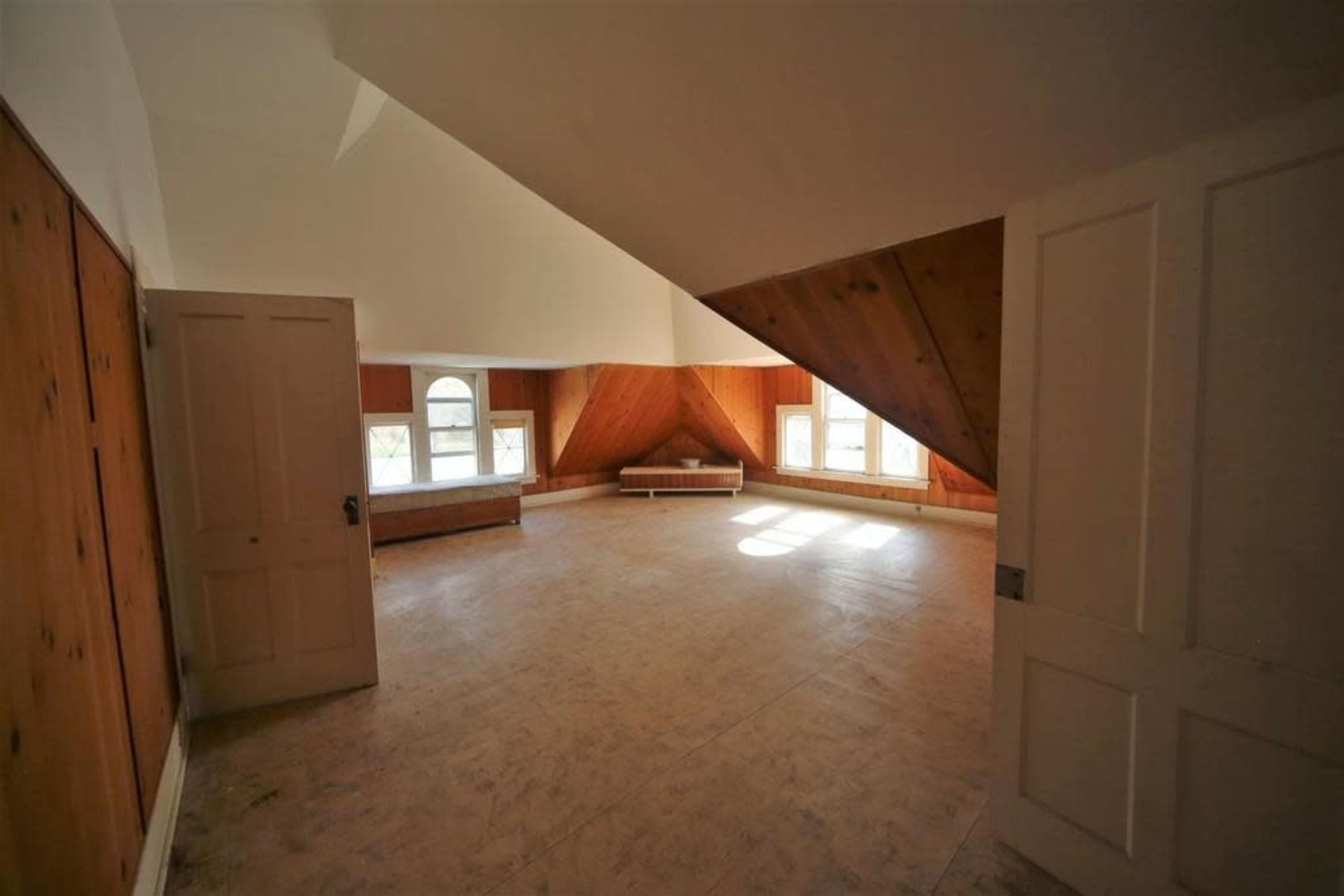 An empty attic room with slanted walls, wooden paneling, and two windows, illuminated by natural light.
