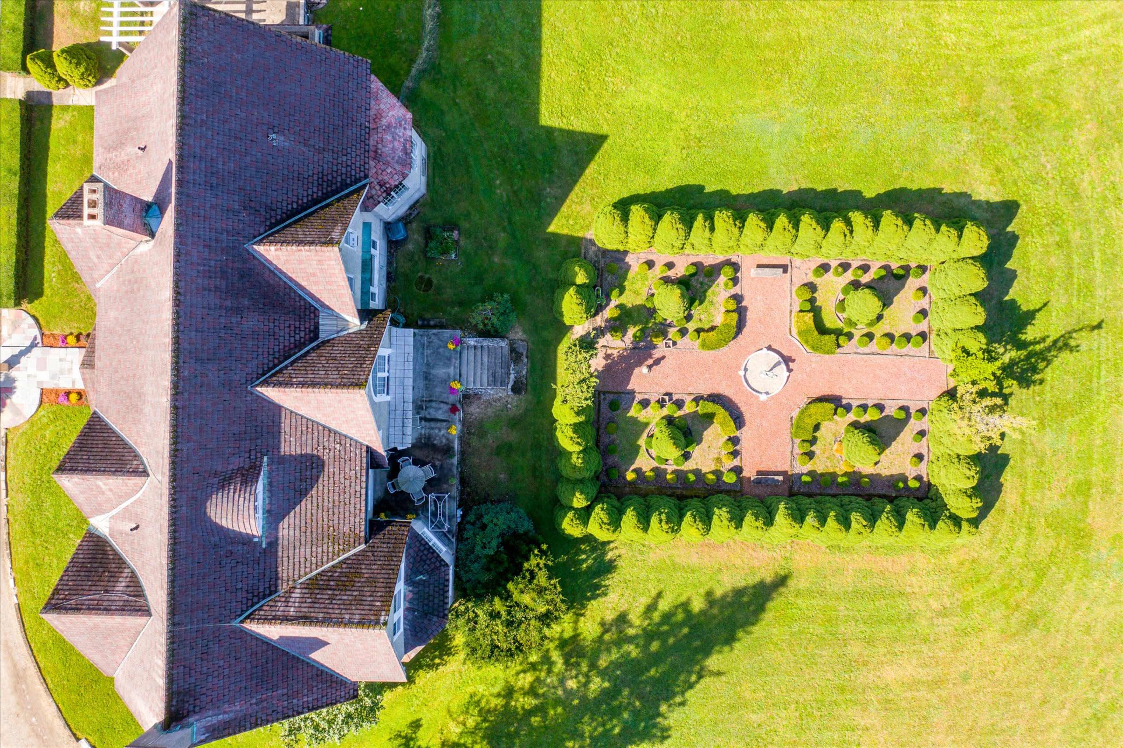 An aerial view shows a large house adjacent to a neatly landscaped garden with symmetrical trees and a central stone feature.