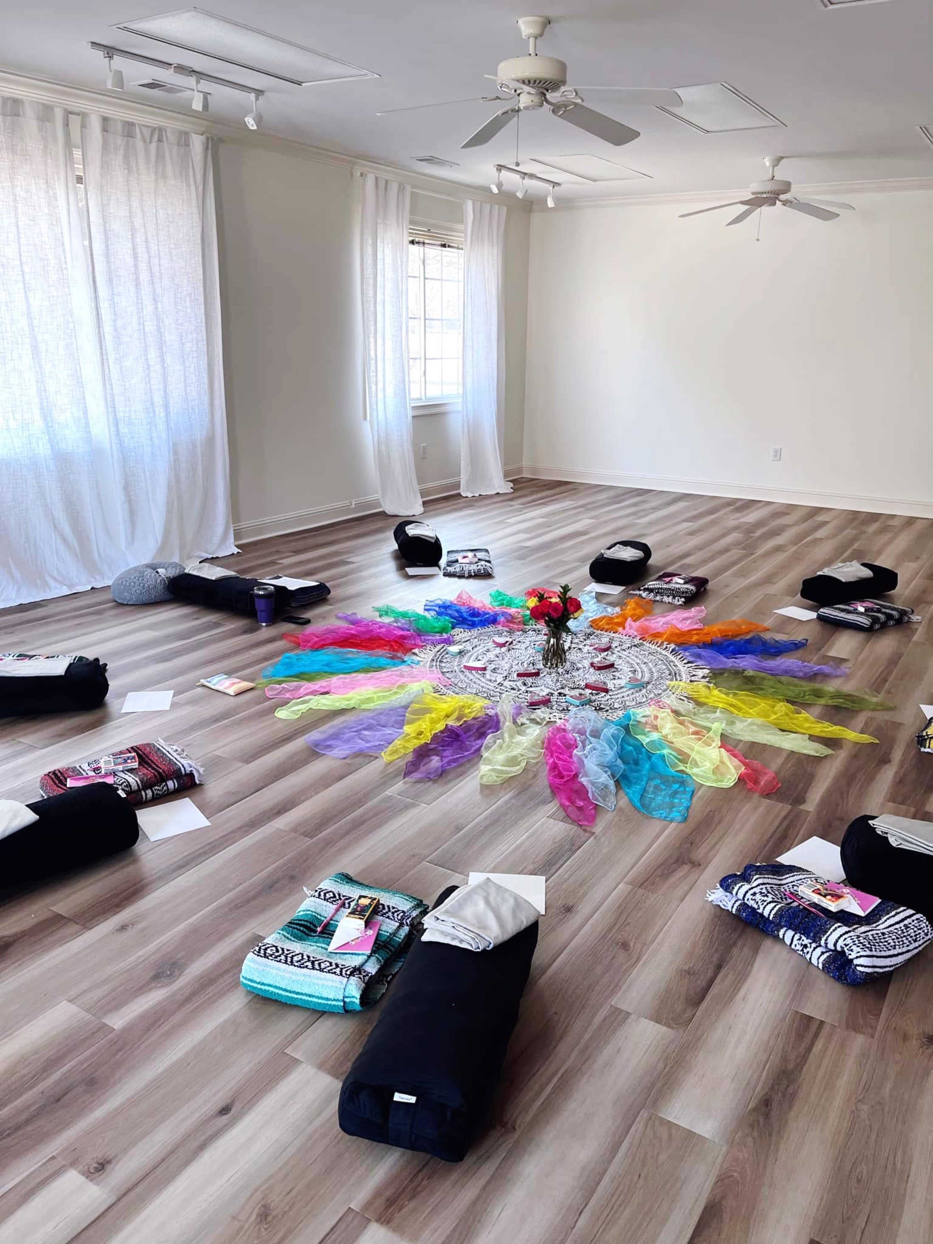 The image shows a spacious, well-lit room with yoga mats arranged in a circle around a colorful mandala made of fabric and flowers at the center.