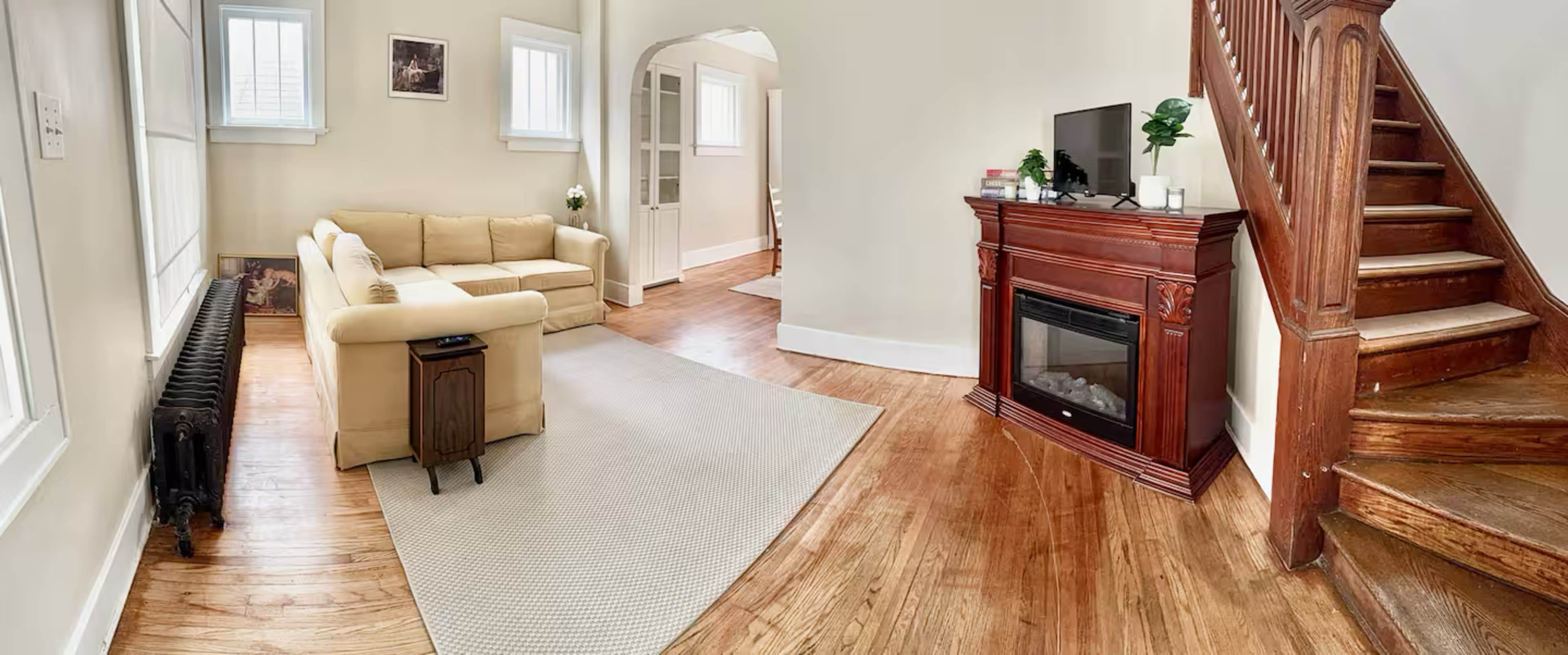 The image shows a bright living room featuring a beige sectional sofa, a wooden staircase, a small table next to the sofa, a fireplace, and hardwood flooring.