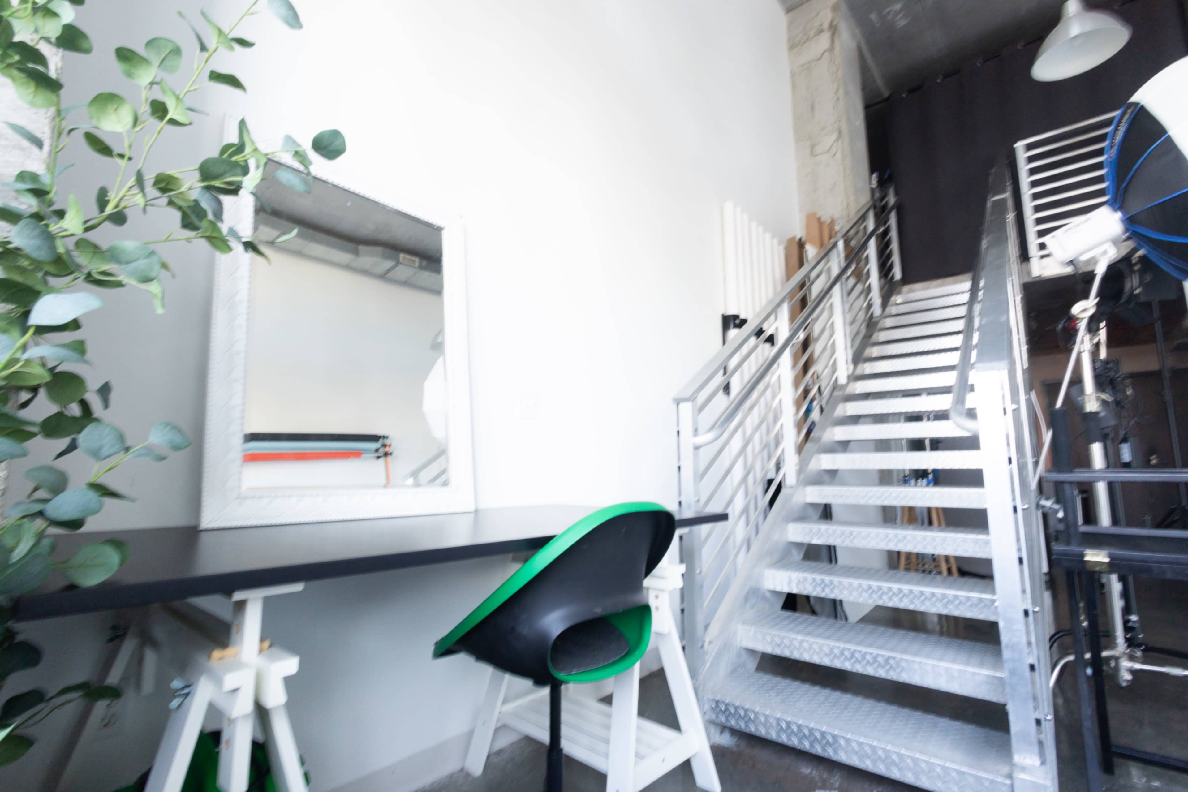The image shows a workspace with a black table and a green chair next to a large mirror, and a metal staircase leading to an upper level.